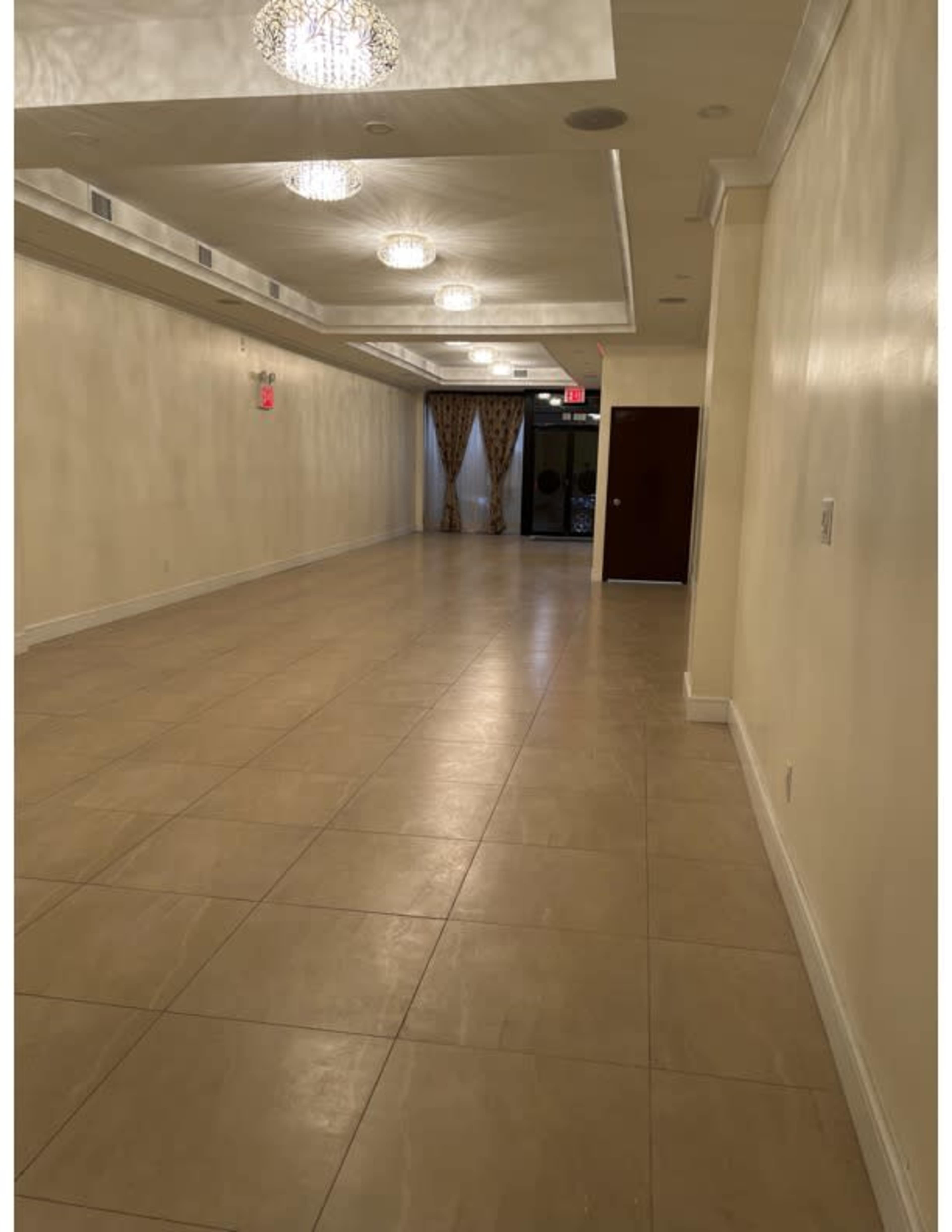 A long, empty hallway with beige tiled flooring, wall sconces, and decorative ceiling lights.