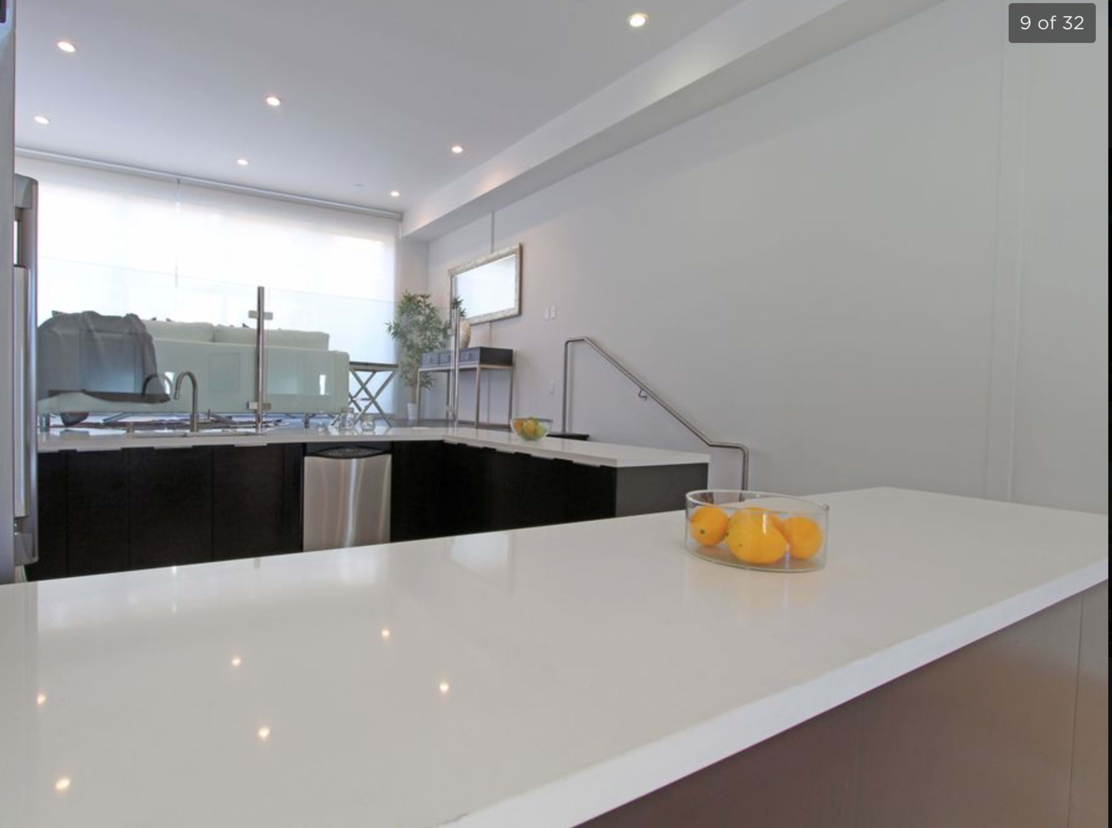 A modern kitchen features a white countertop with a bowl of lemons, stainless steel appliances, and a sliding door that leads to a living area.