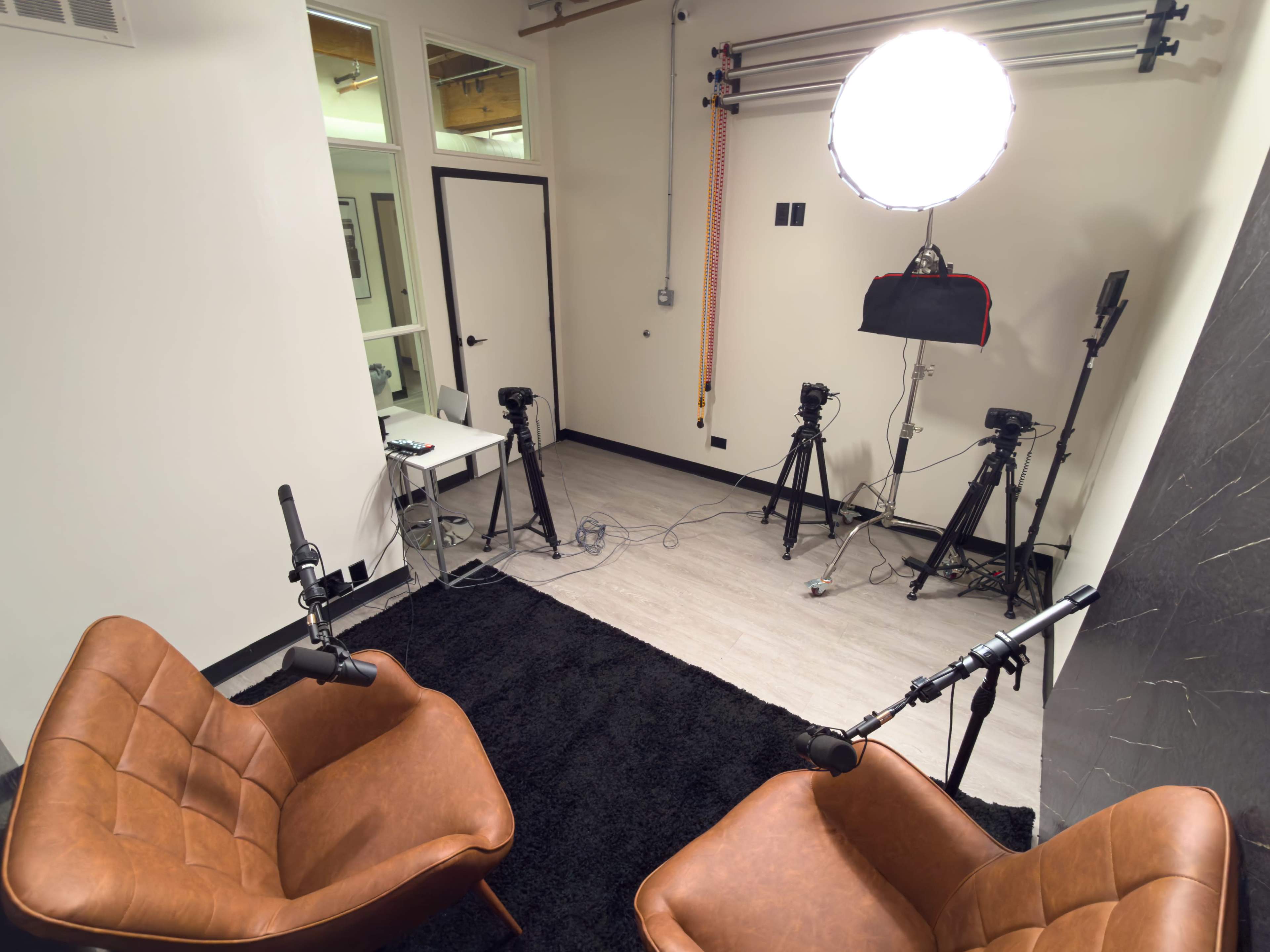 The image shows a small studio setup with two leather chairs facing each other, surrounded by multiple cameras and a bright circular light.