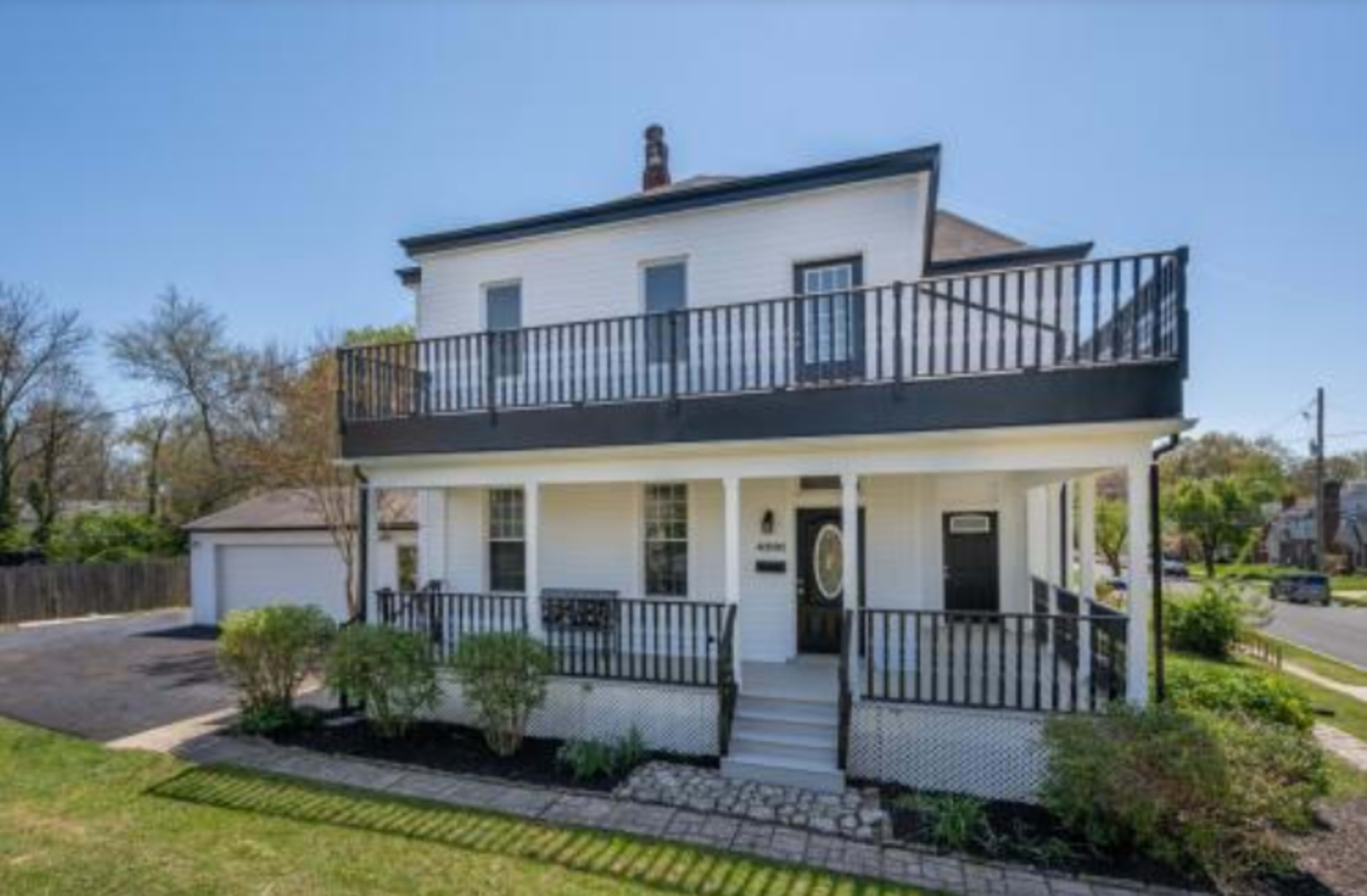 A two-story white house with a black balcony and a front porch, situated on a well-maintained lawn next to a paved driveway.