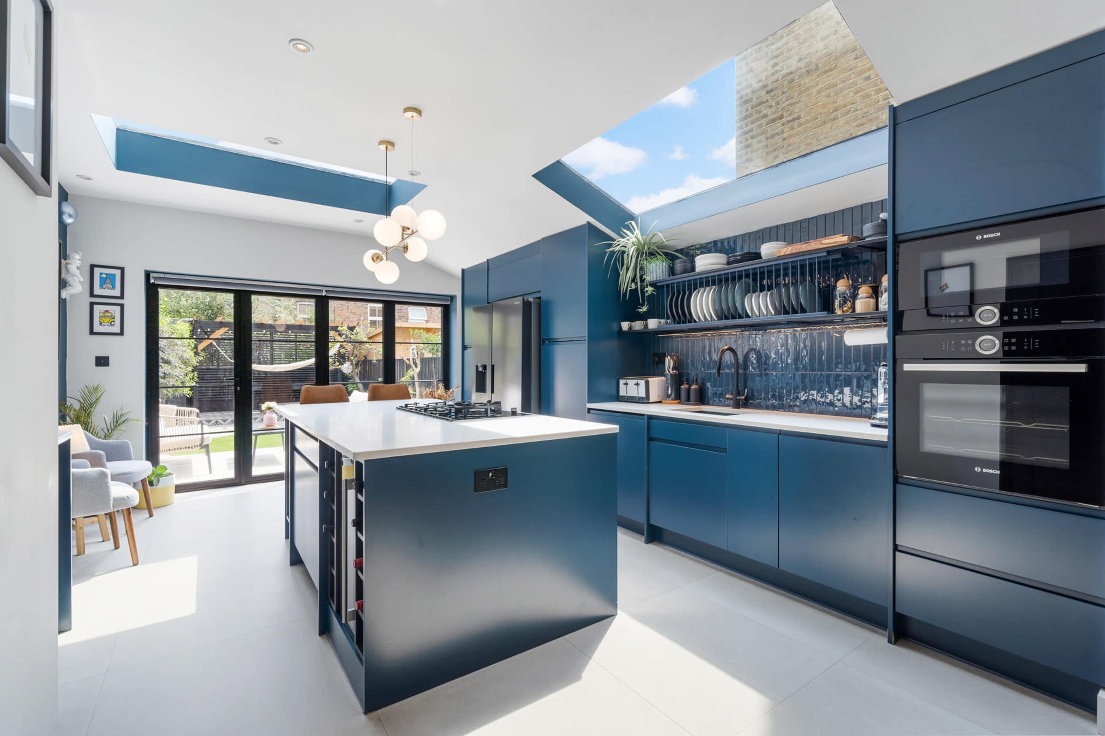 The image shows a modern kitchen with blue cabinetry, a large island with a cooktop, and a skylight that brightens the space.
