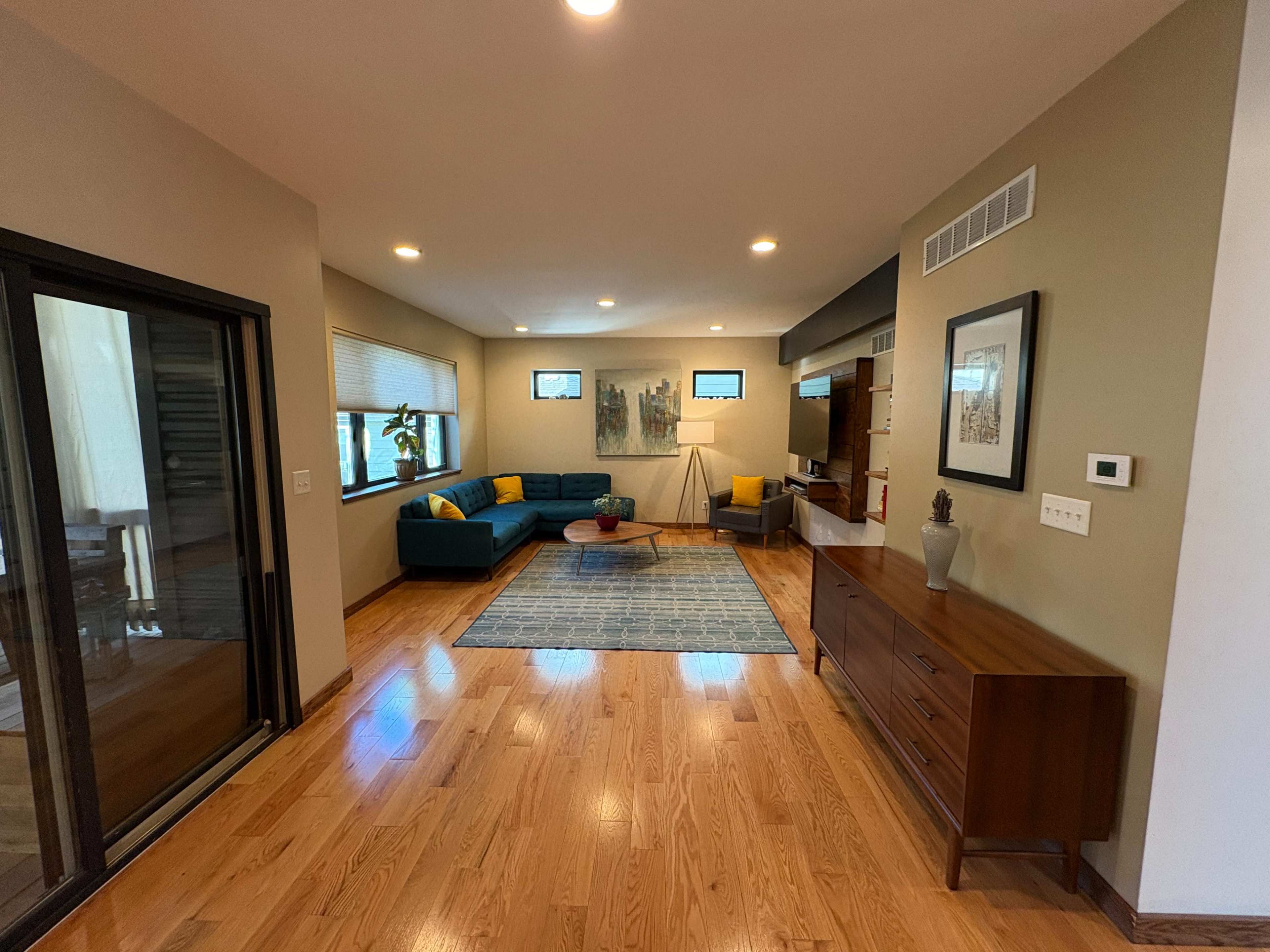 The image shows a modern living room with a green sofa, a wooden cabinet, and large windows allowing natural light.