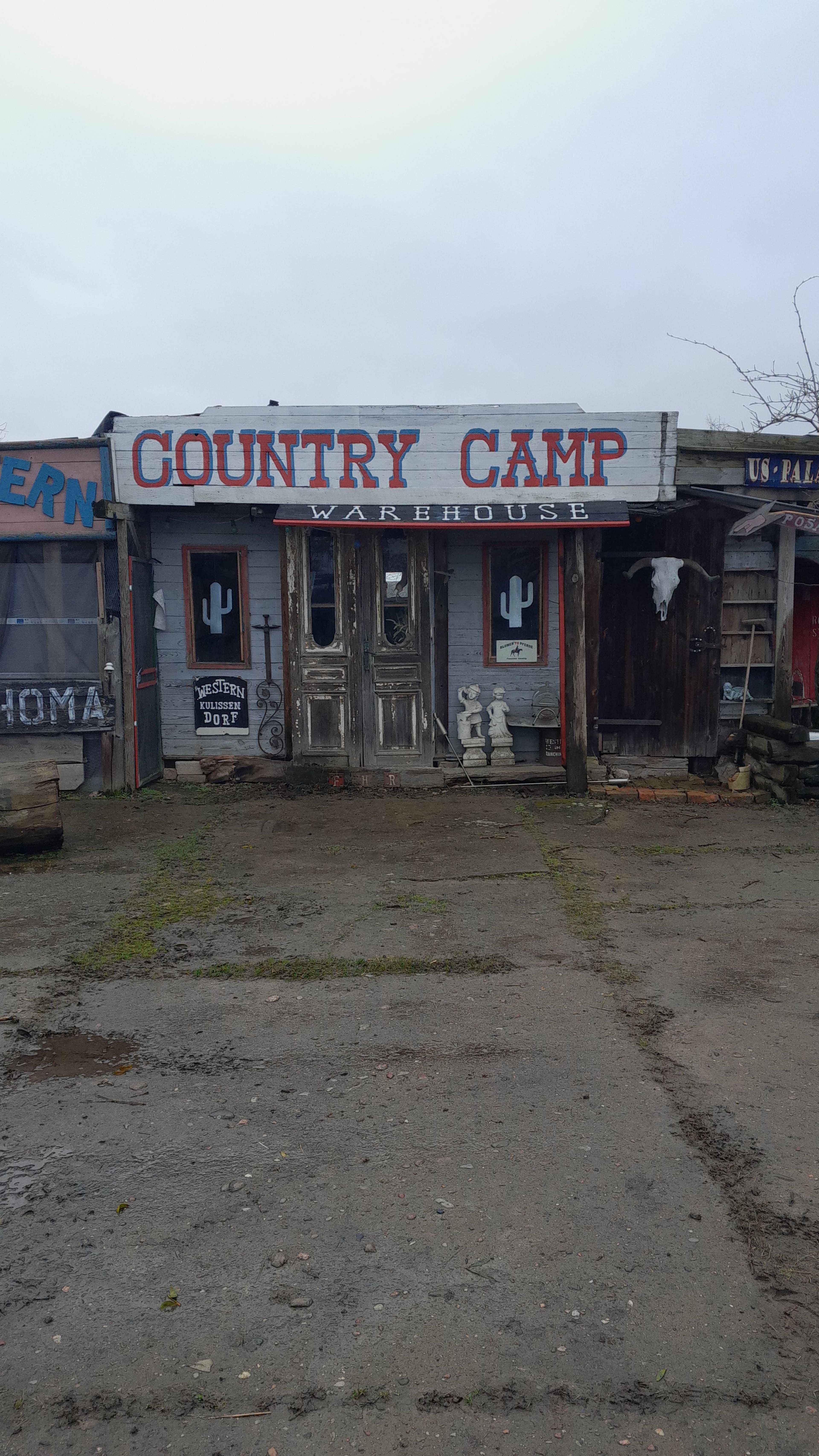 The image shows an old, rustic building with a sign that reads "Country Camp Warehouse" and two doors in the front.