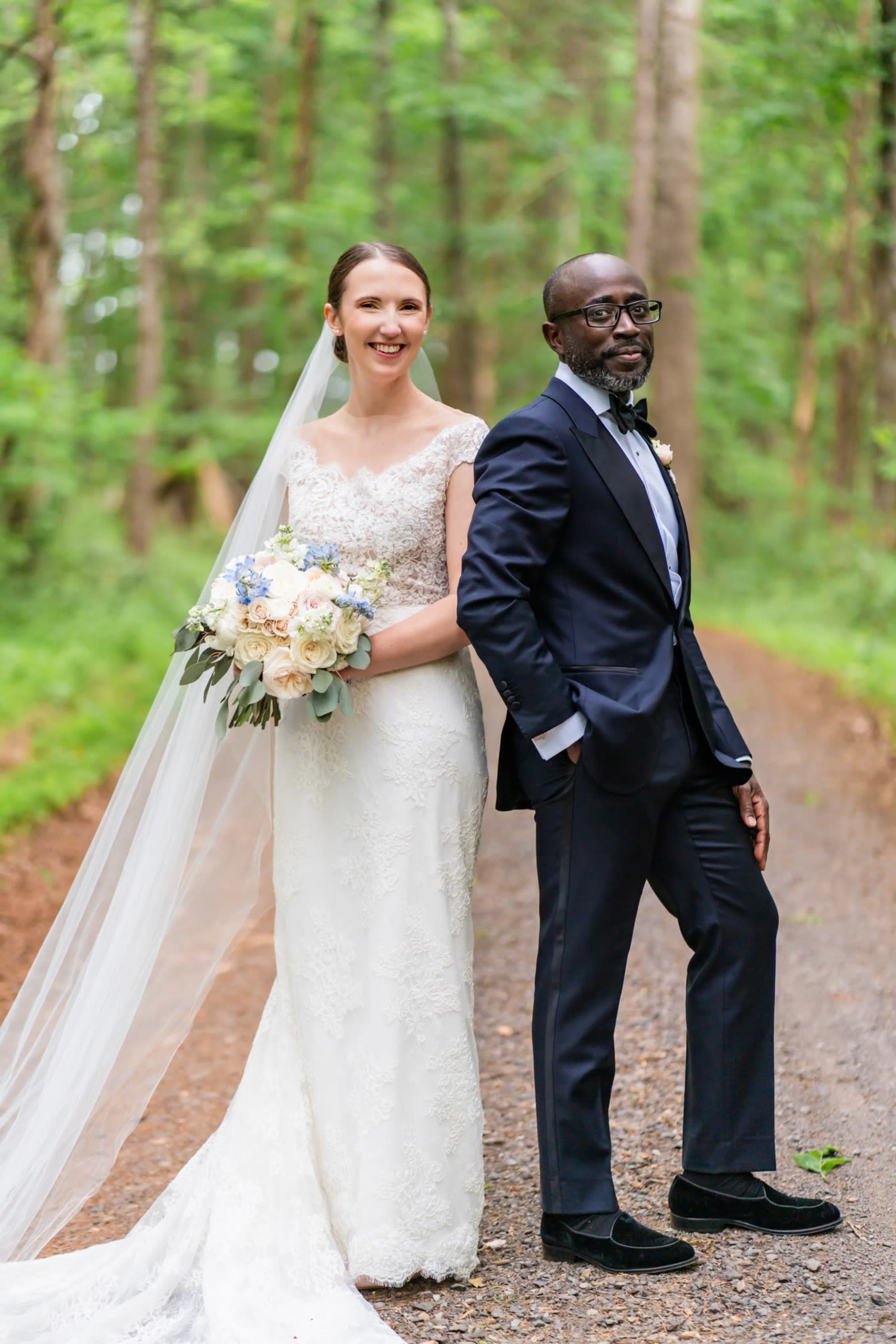 A bride in a lace wedding gown and a groom in a suit pose together on a gravel path surrounded by trees.