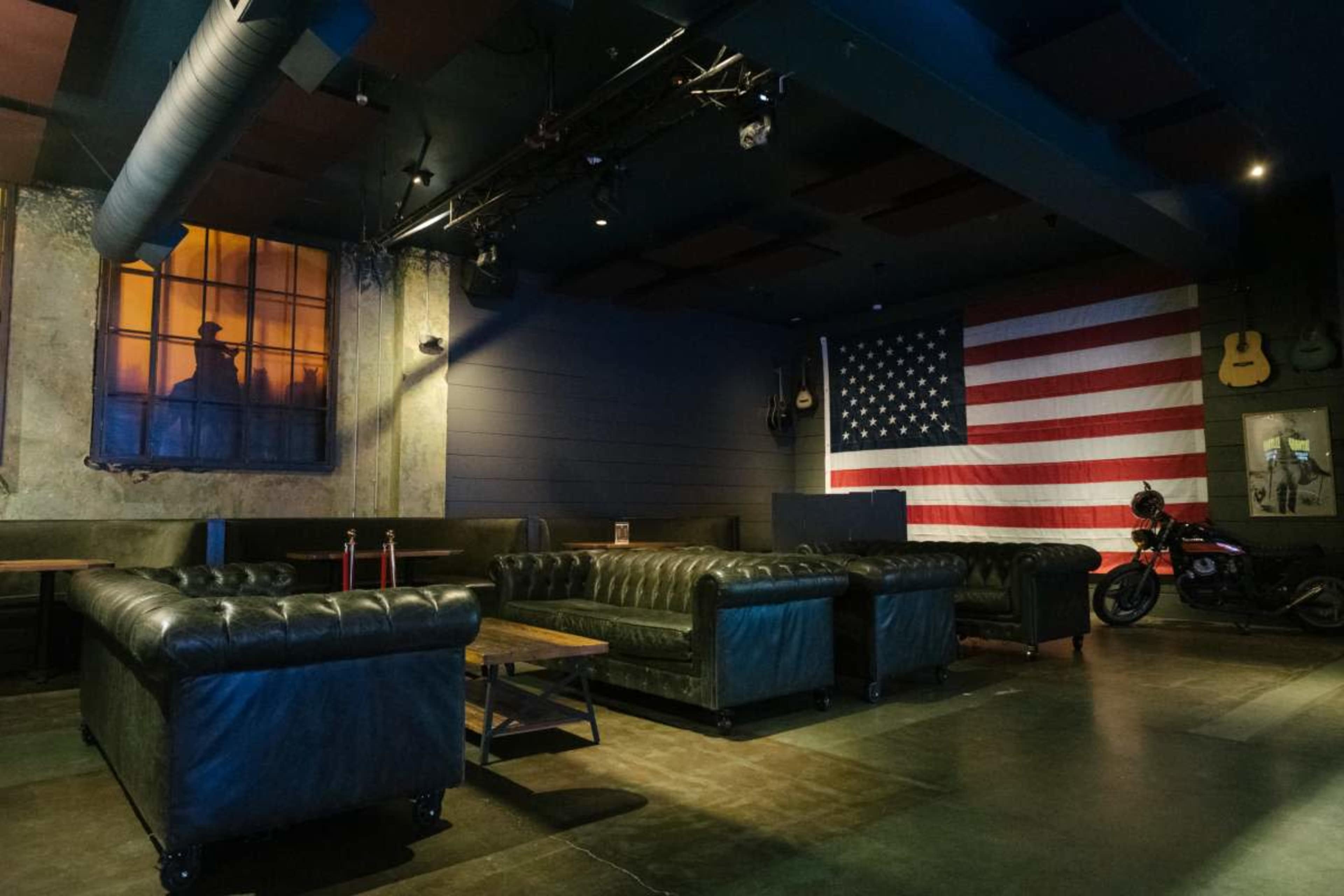 The image shows a dimly lit lounge area featuring black leather couches, an American flag on the wall, and a vintage motorcycle parked nearby.