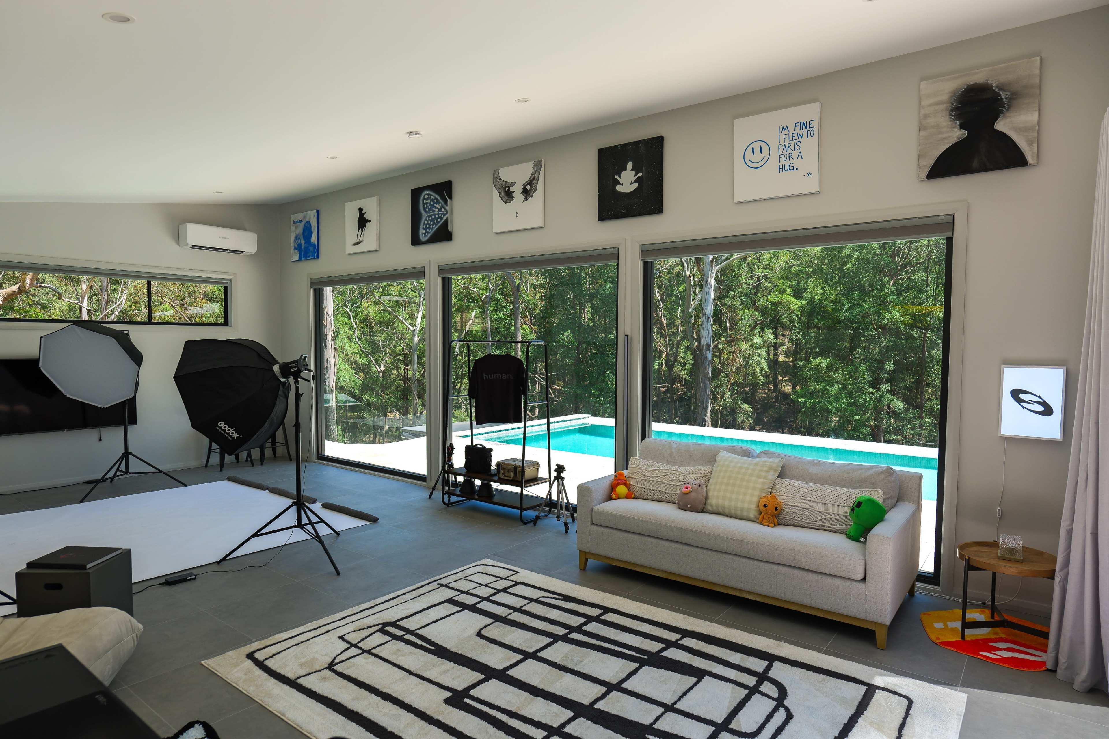 A modern living room features large windows overlooking a pool and lush greenery, with contemporary artwork on the walls and a sofa arranged with colorful cushions.