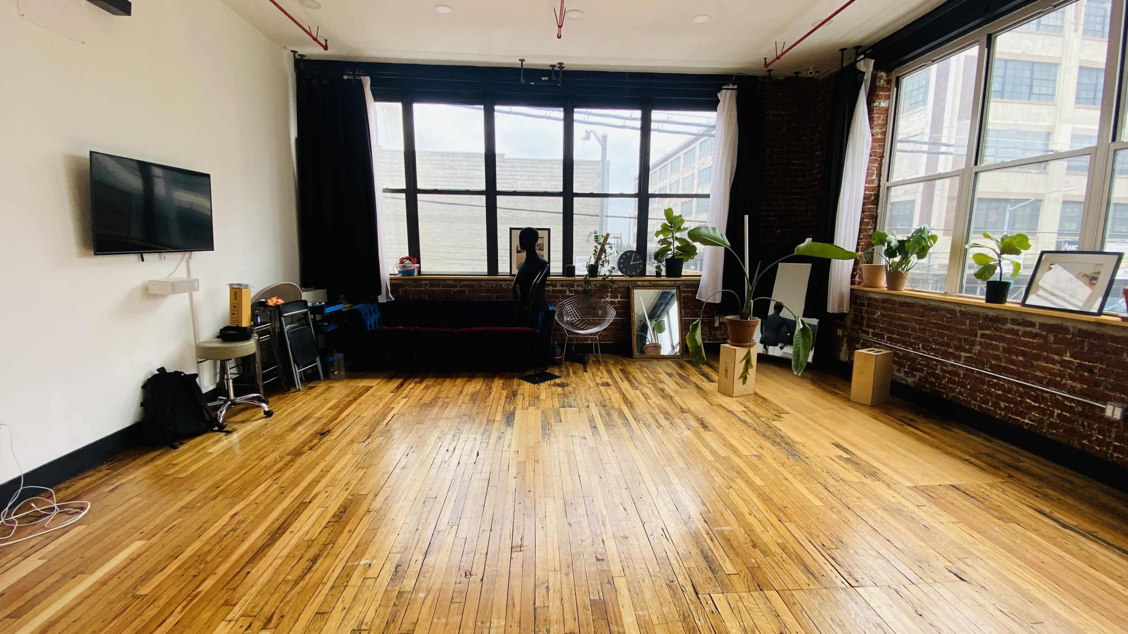 The image shows a spacious room with wooden floors, large windows, plants, a sofa, and various pieces of furniture.