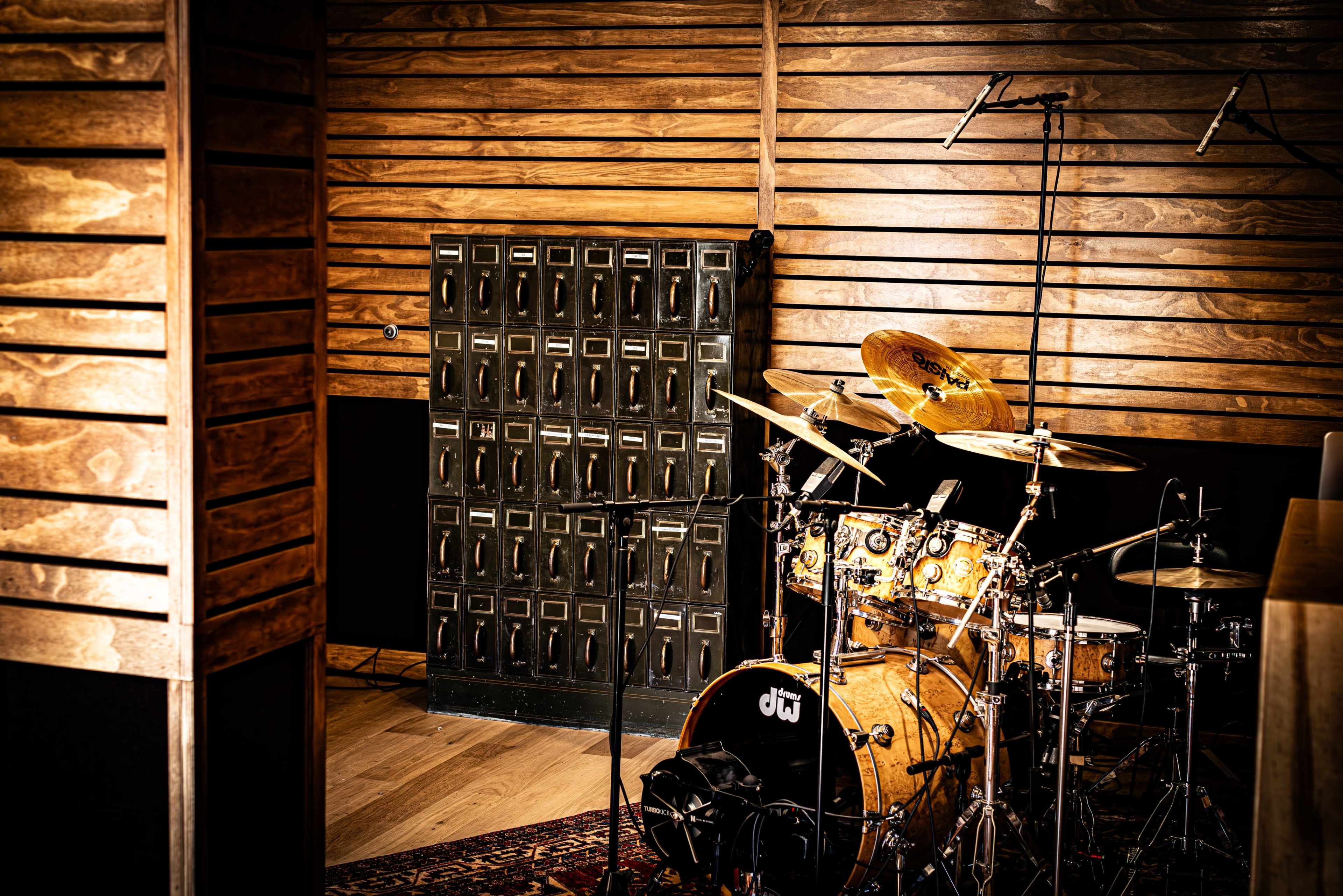 The image shows a drum set positioned in a wooden-paneled room beside a large, black storage cabinet.