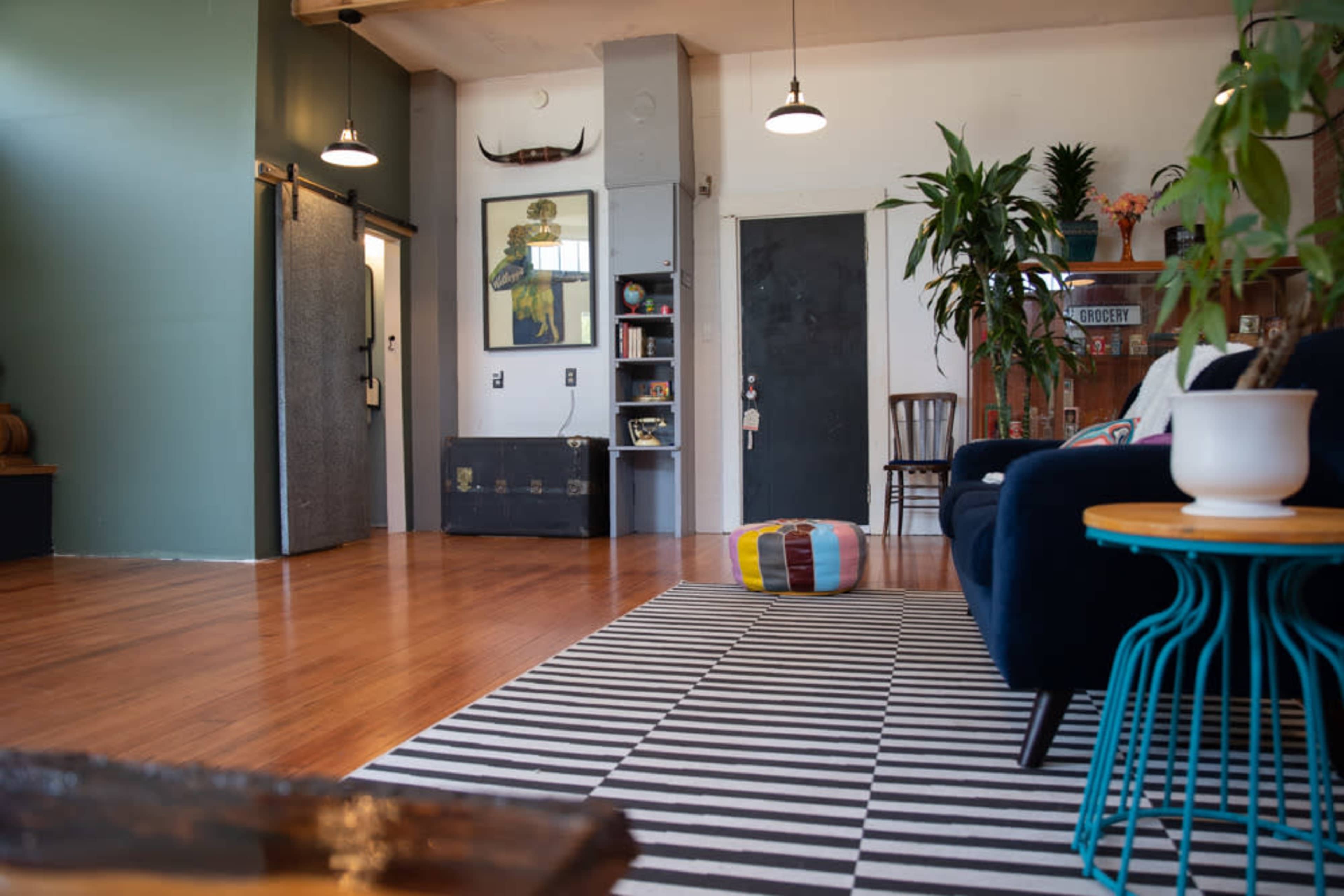 The image shows a spacious, modern living room with wooden flooring, a striped area rug, and a mix of plants and furniture.