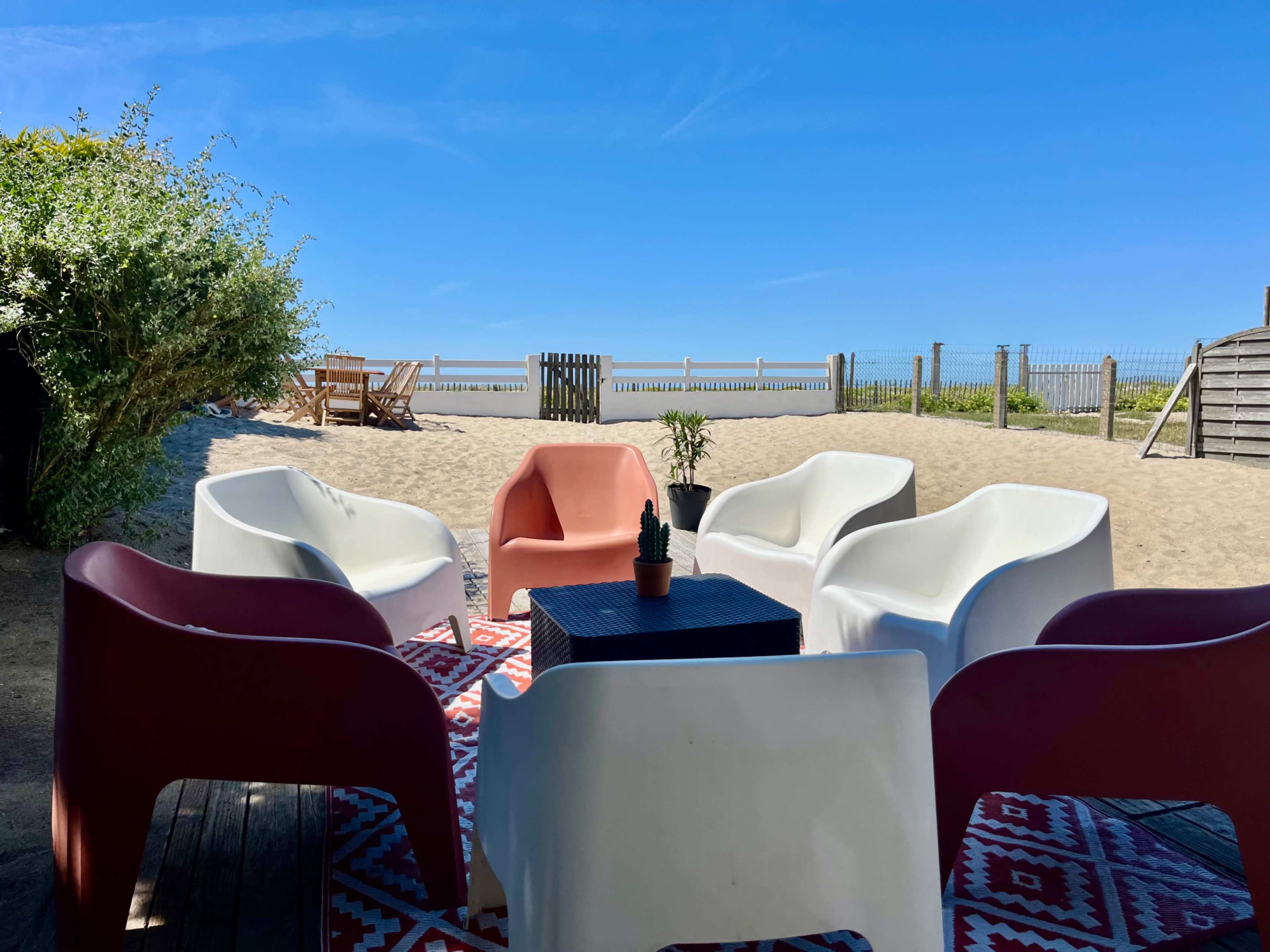A circular arrangement of six colorful chairs surrounds a small table on a sandy beach with a clear blue sky in the background.