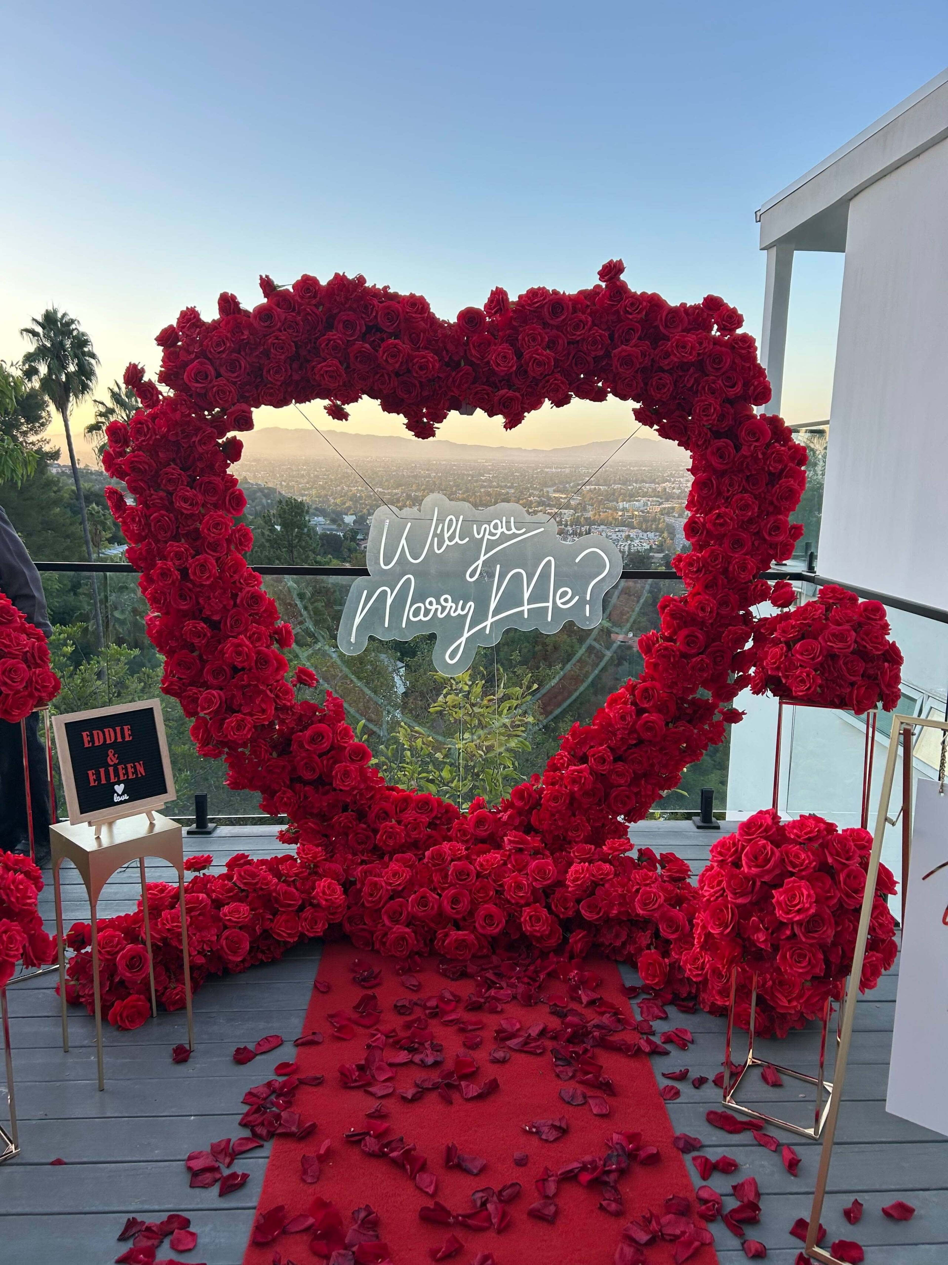 The image shows a large heart-shaped arrangement of red roses with the words "Will you Marry Me?" displayed in white, set against a scenic backdrop.