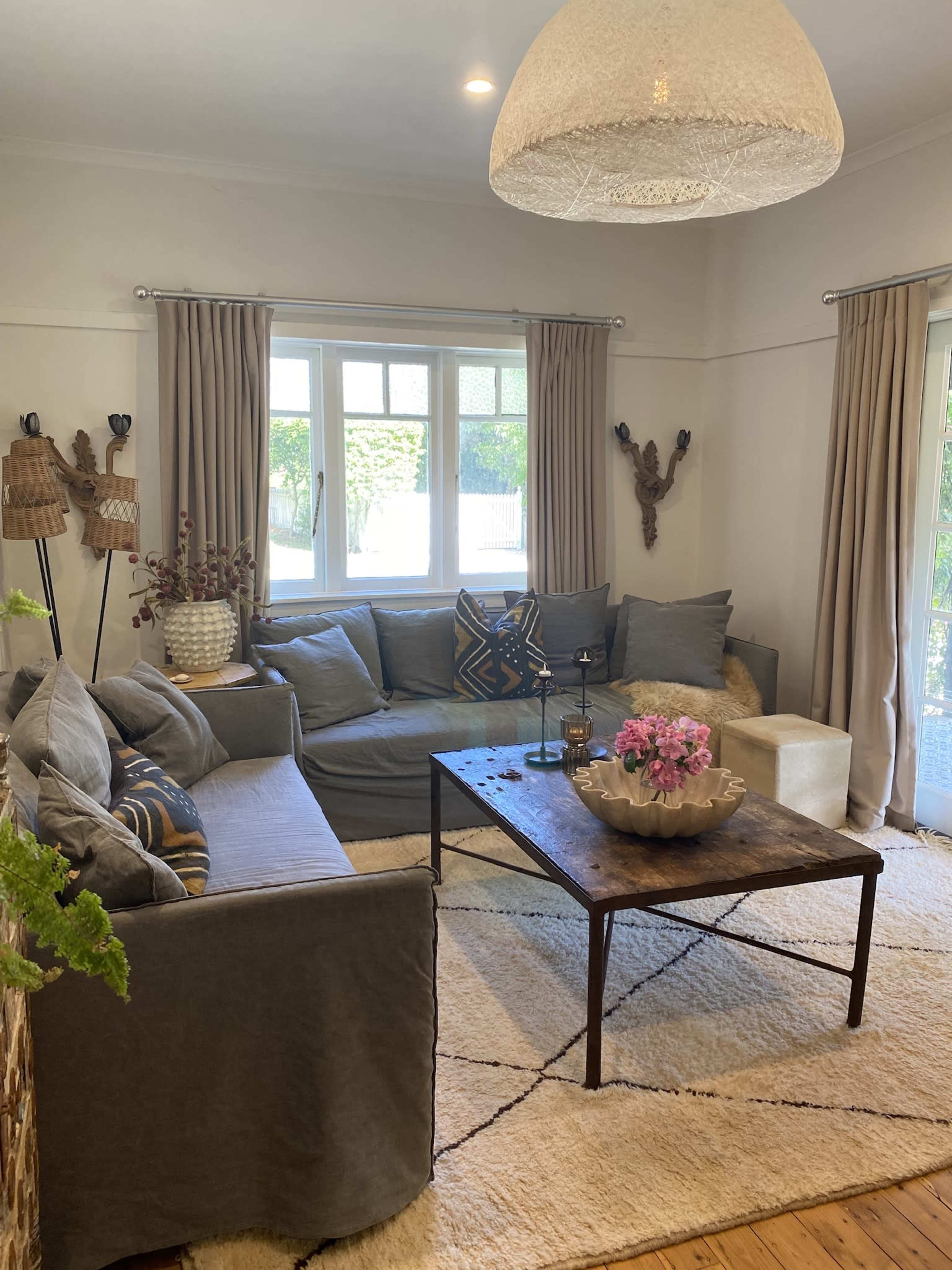 The image shows a modern living room featuring a large gray sectional sofa, a wooden coffee table with a bowl of flowers, and large windows dressed with light curtains, with a plush rug on the wooden floor.