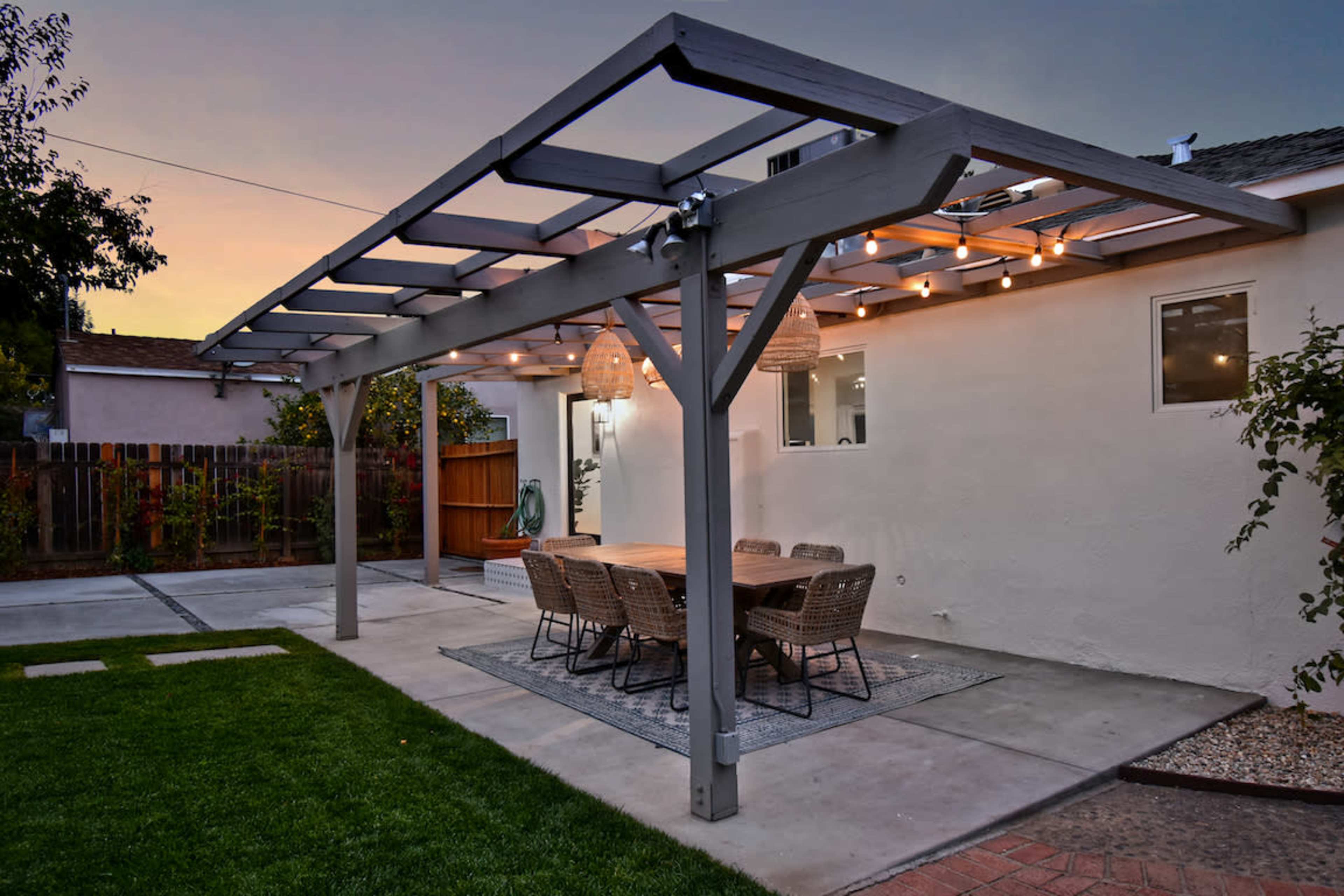 The image shows a patio area with a wooden pergola, a large dining table surrounded by wicker chairs, and decorative lights strung above in a residential backyard.
