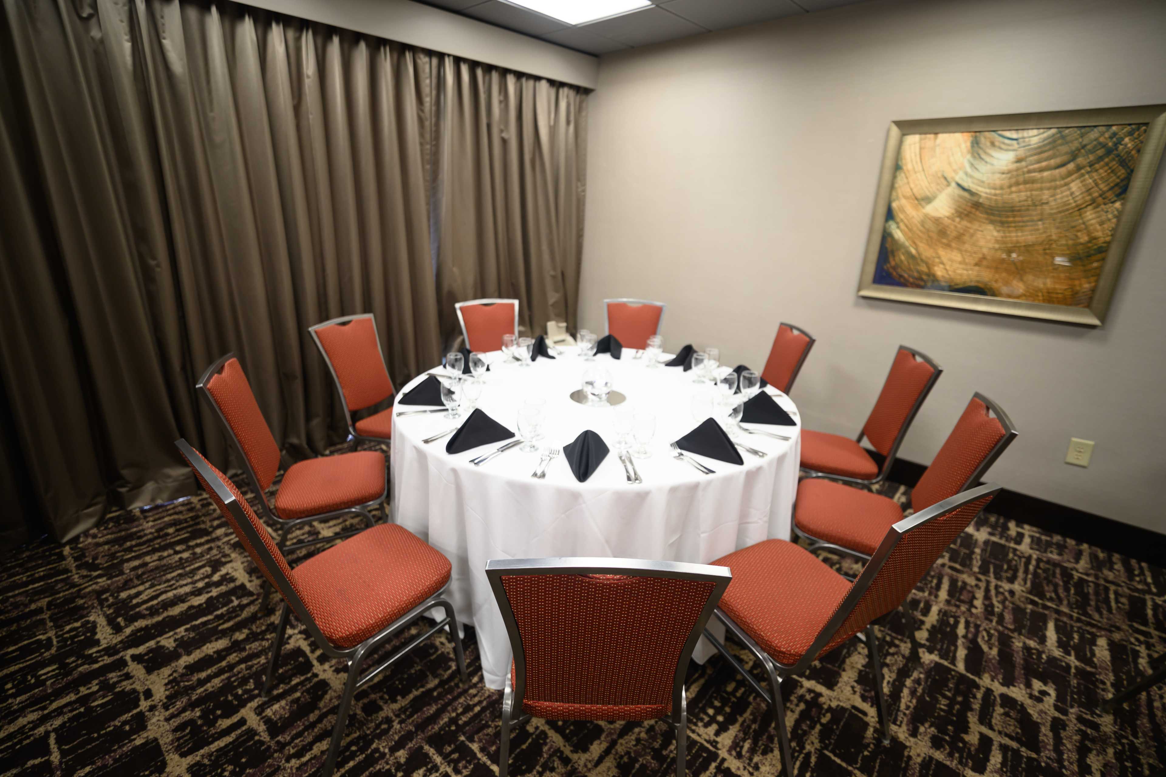 A round dining table is set for ten with white tablecloth, black napkins, and glassware, surrounded by red upholstered chairs in a well-lit room.