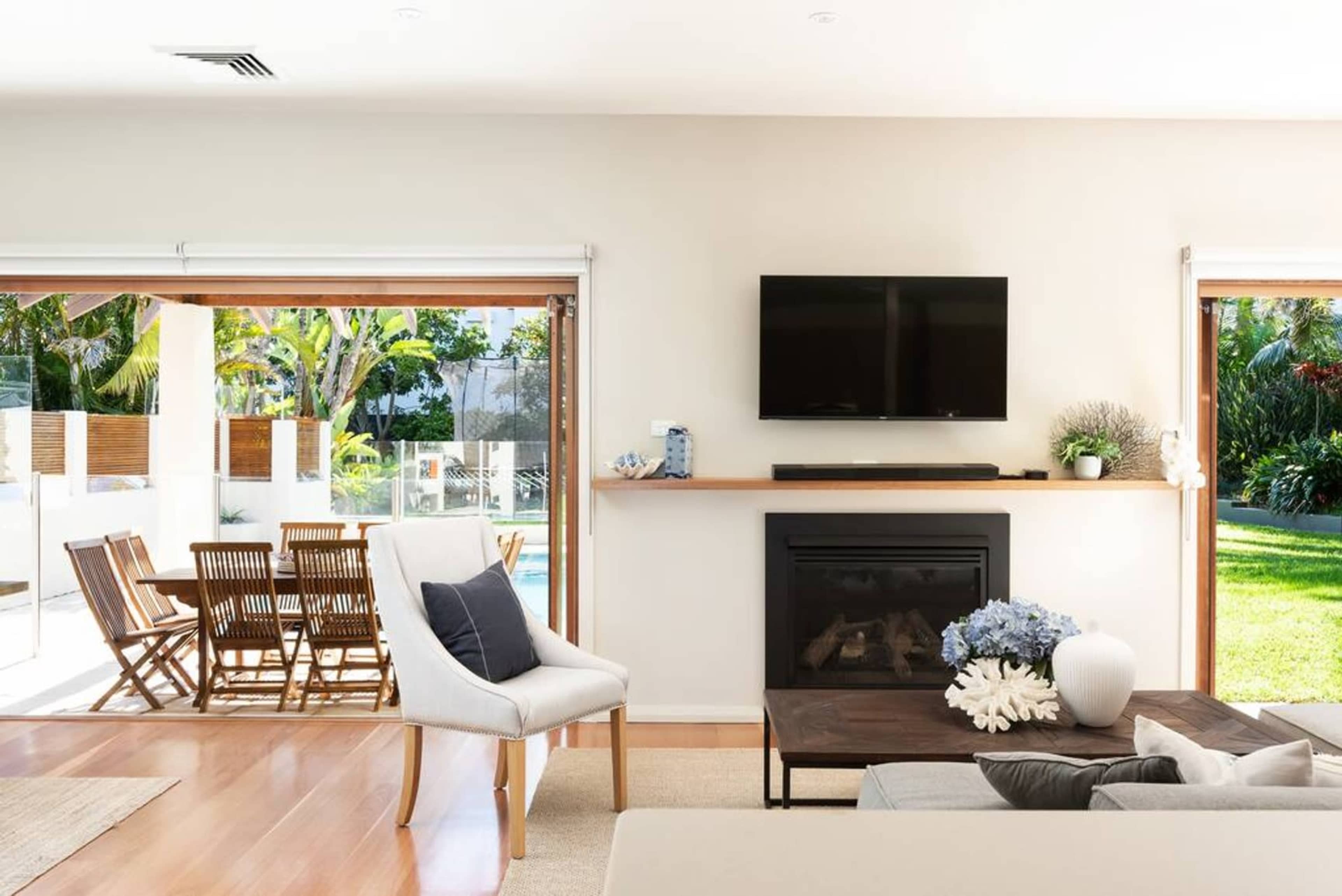 A modern living room featuring a fireplace, a television mounted on the wall, and large windows overlooking a patio with outdoor seating and a pool.