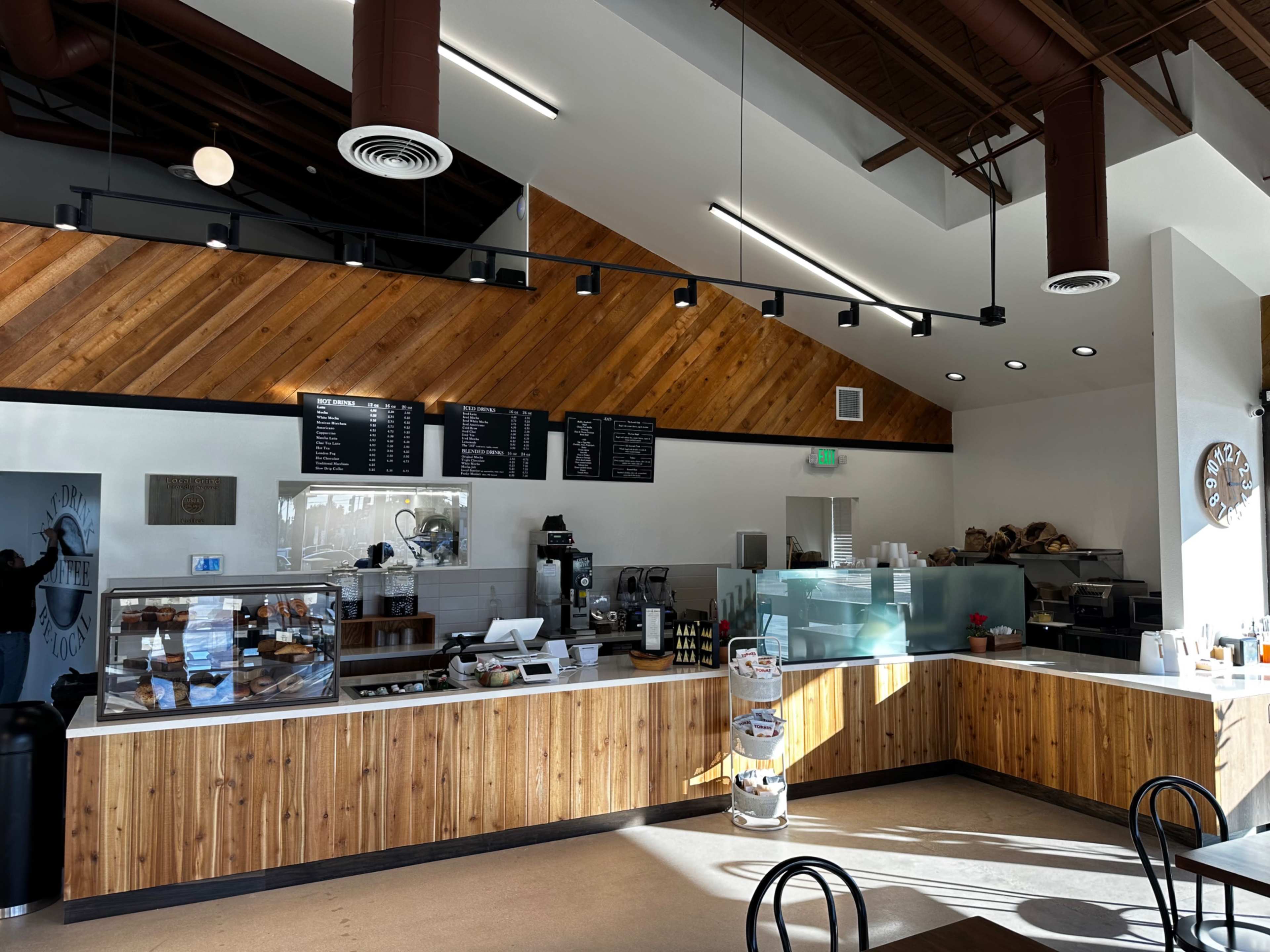 A modern café with a wooden counter, menu boards, and an open layout featuring a coffee preparation area and display case for pastries.