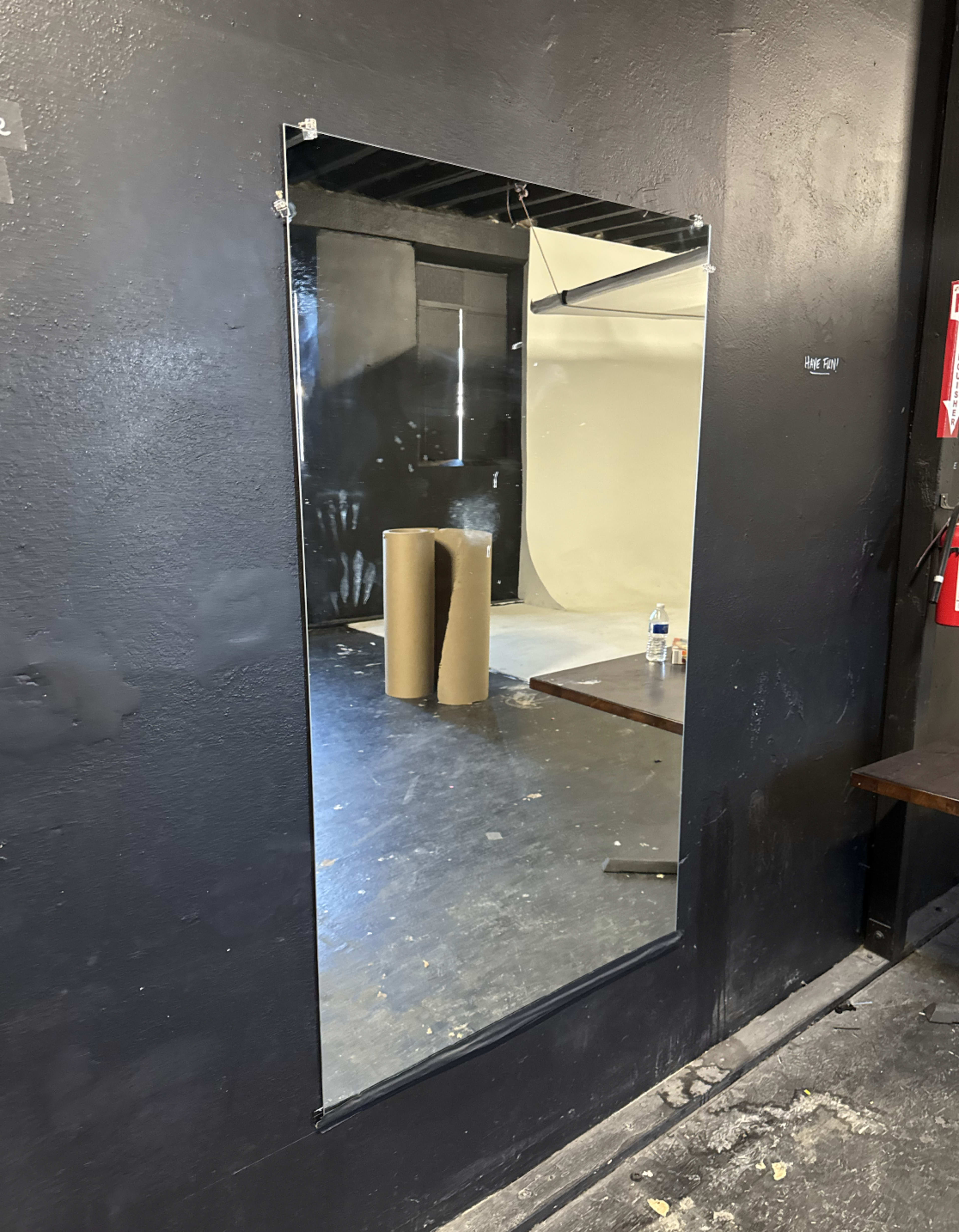 A large, rectangular mirror is mounted on a black wall in a studio space, reflecting a backdrop and some equipment.