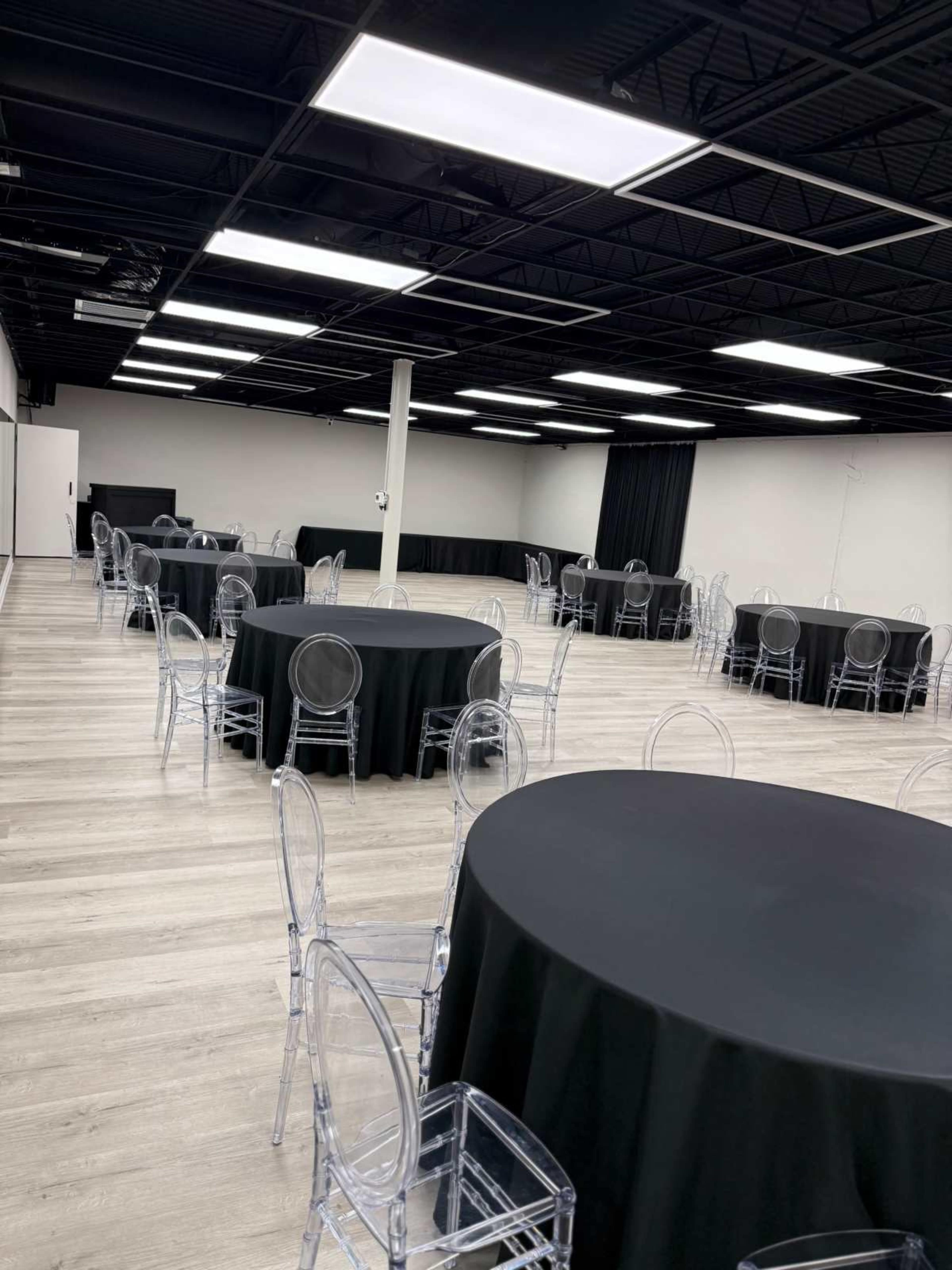 The image shows an empty event space with several round tables covered in black cloth and transparent chairs arranged throughout the room.