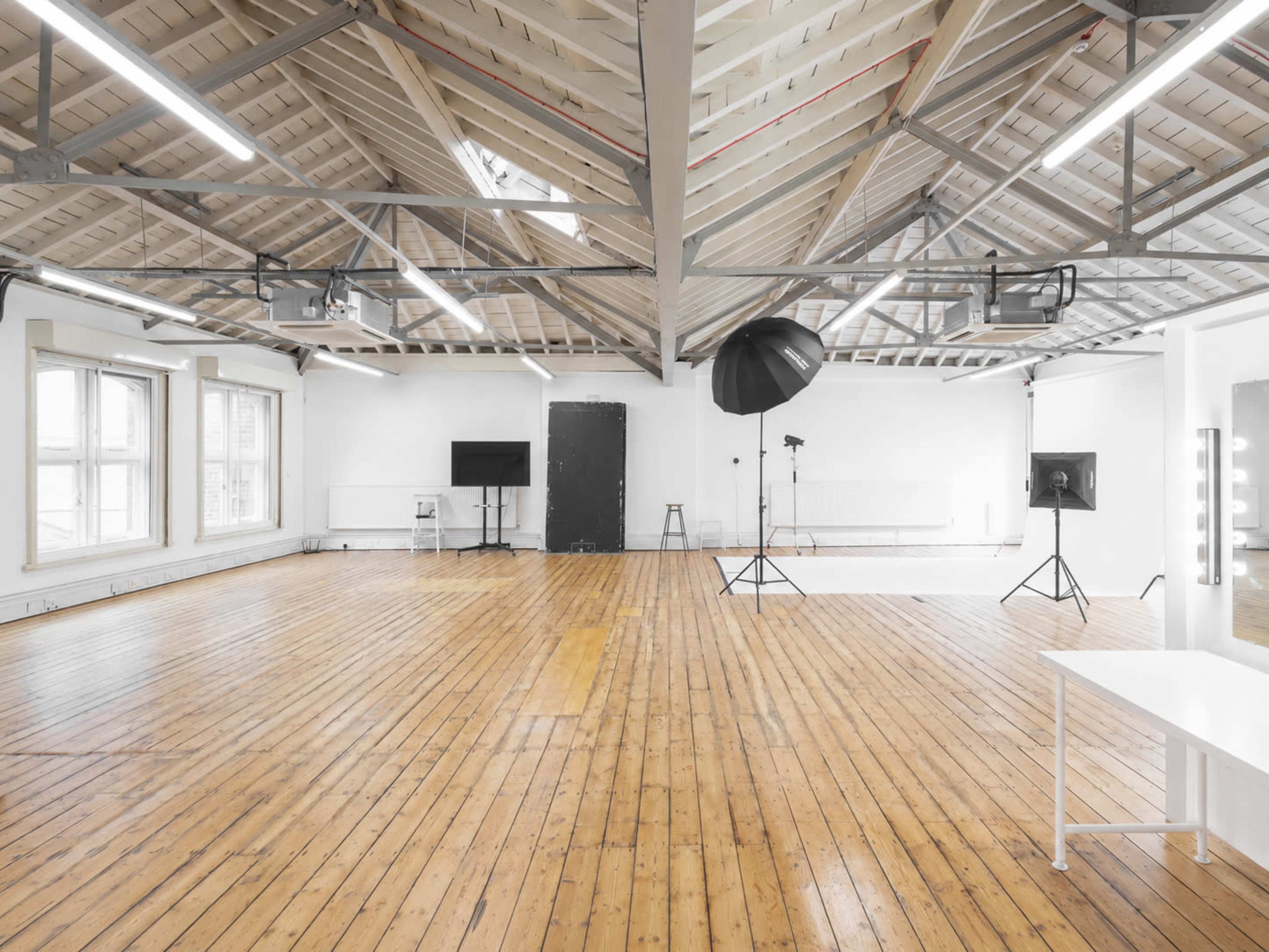 The image depicts a spacious, well-lit photography studio featuring wooden floors, large windows, and multiple lighting setups.