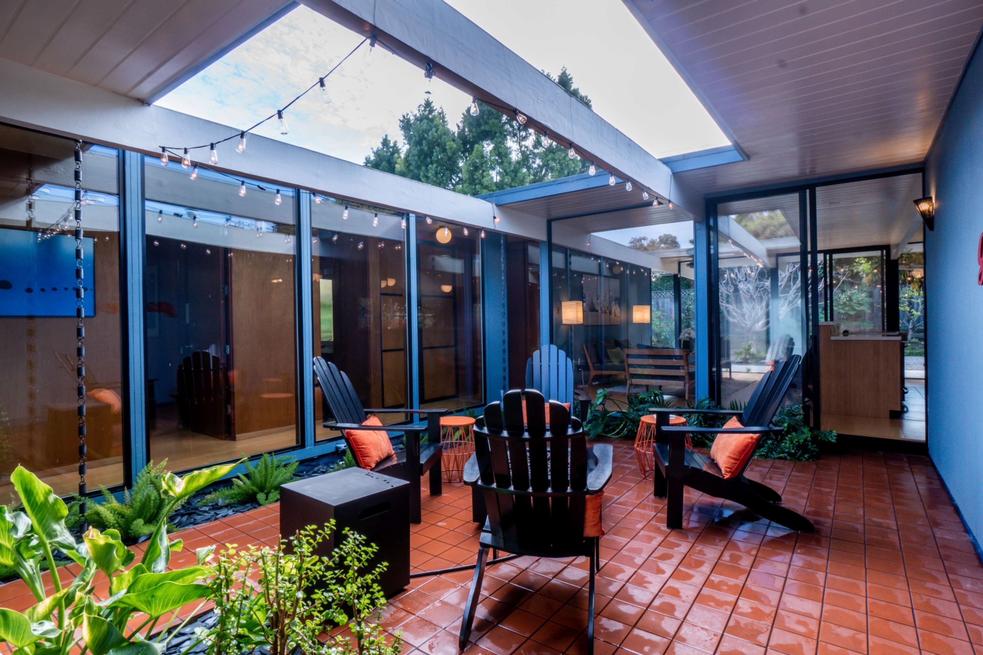 The image shows a modern courtyard featuring four wooden chairs arranged around a small table, with decorative string lights overhead and glass walls on either side.