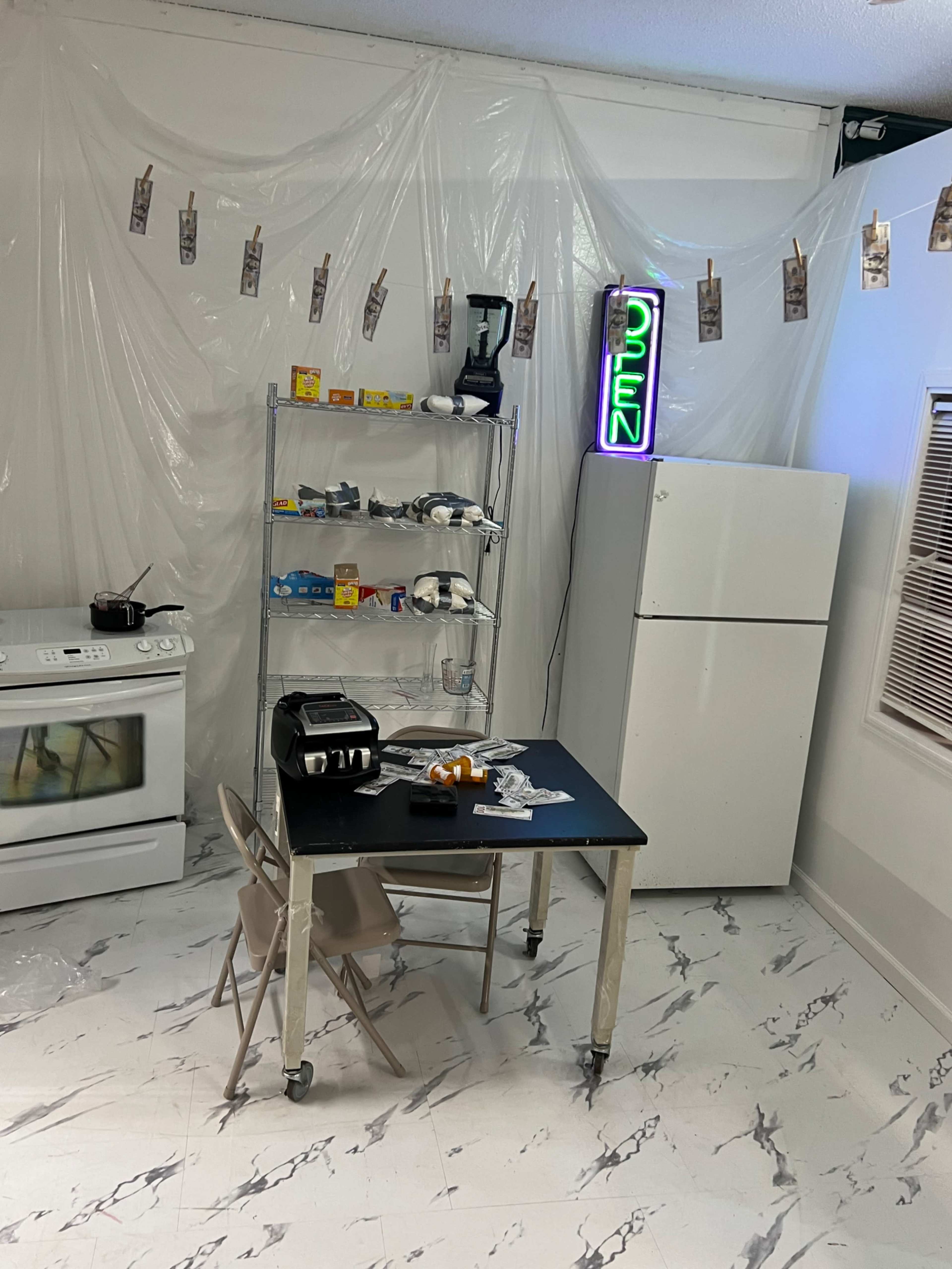 A kitchen features a plastic-covered wall, a white stove, a fridge with a neon sign, a metal shelf stocked with food items, and a small table with a few scattered objects.