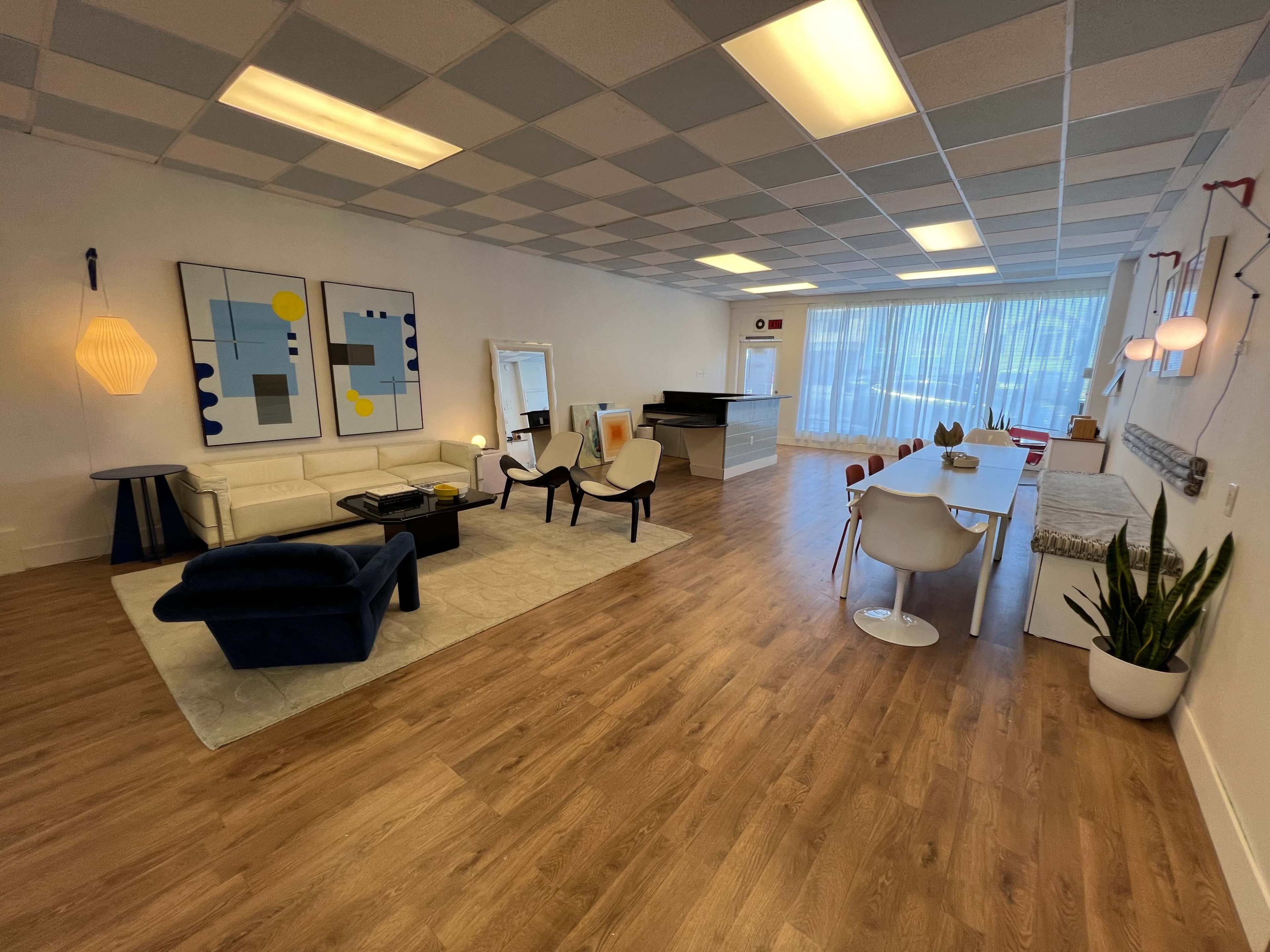 The image shows a spacious, well-lit room with a modern living area featuring a white sofa, chairs, and a dining table, along with large windows and artwork on the walls.