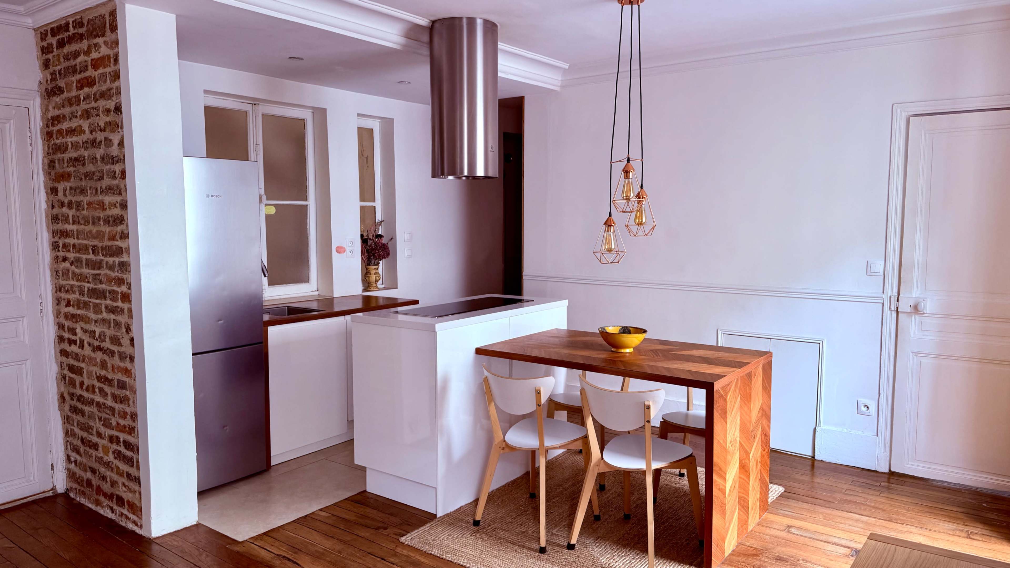 The image shows a modern kitchen and dining area with a wooden table and two chairs, a stainless steel refrigerator, and contemporary light fixtures.