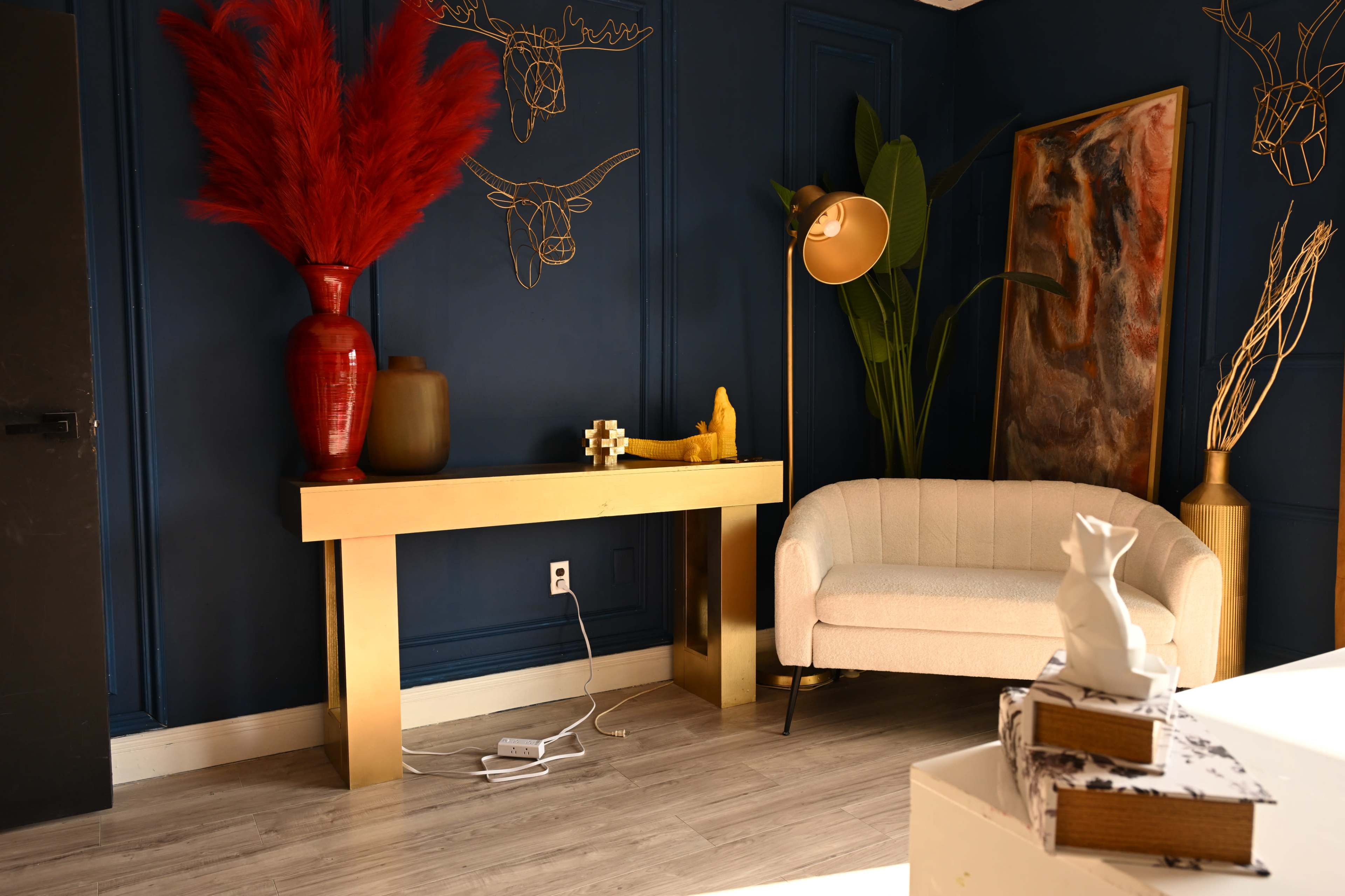 The image shows a modern living space featuring a gold console table, a white sofa, and decorative elements including plants and artwork against a dark blue wall.