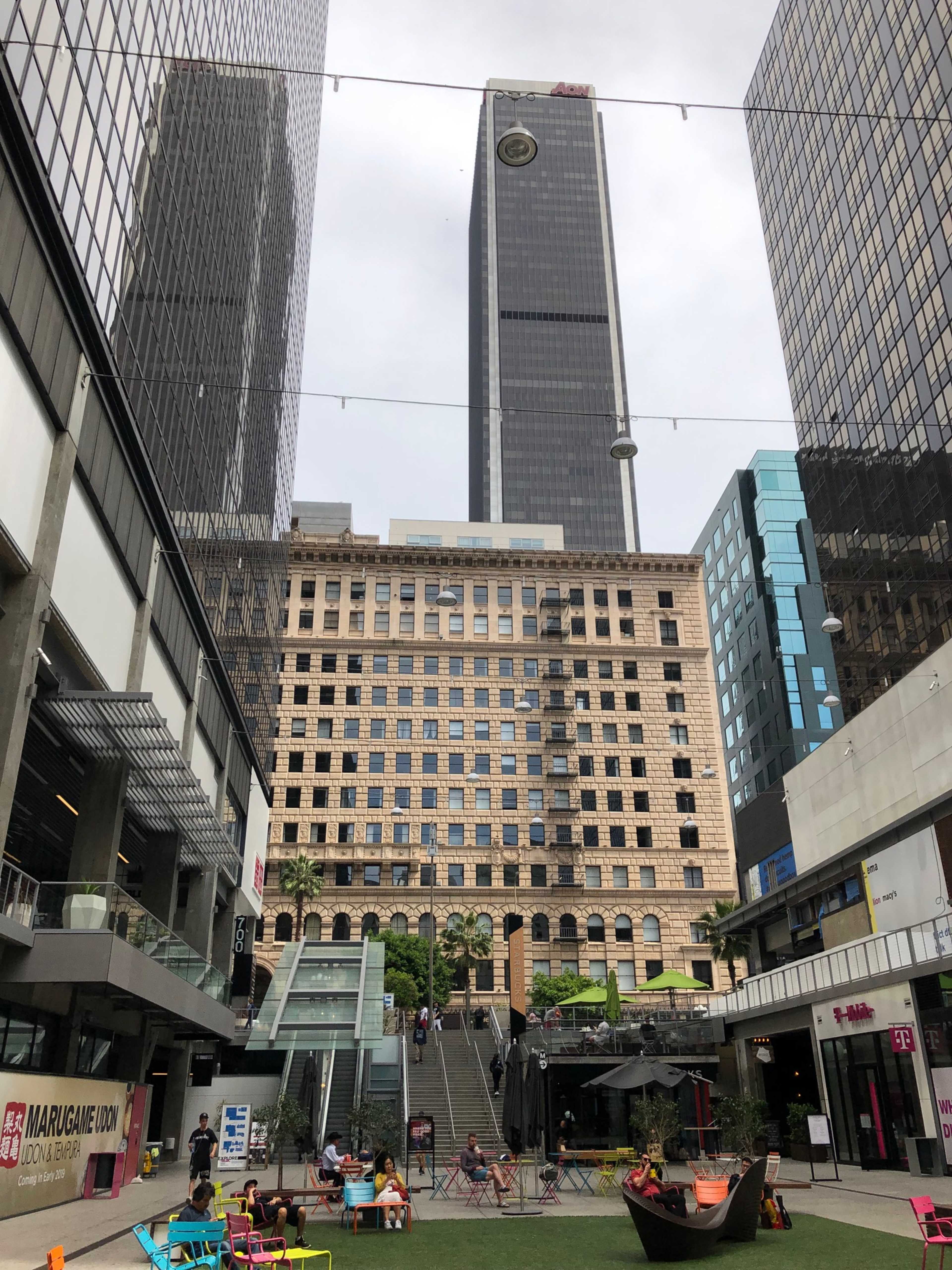 The image shows a modern urban plaza surrounded by tall buildings, with a historical structure in the center and outdoor seating areas.