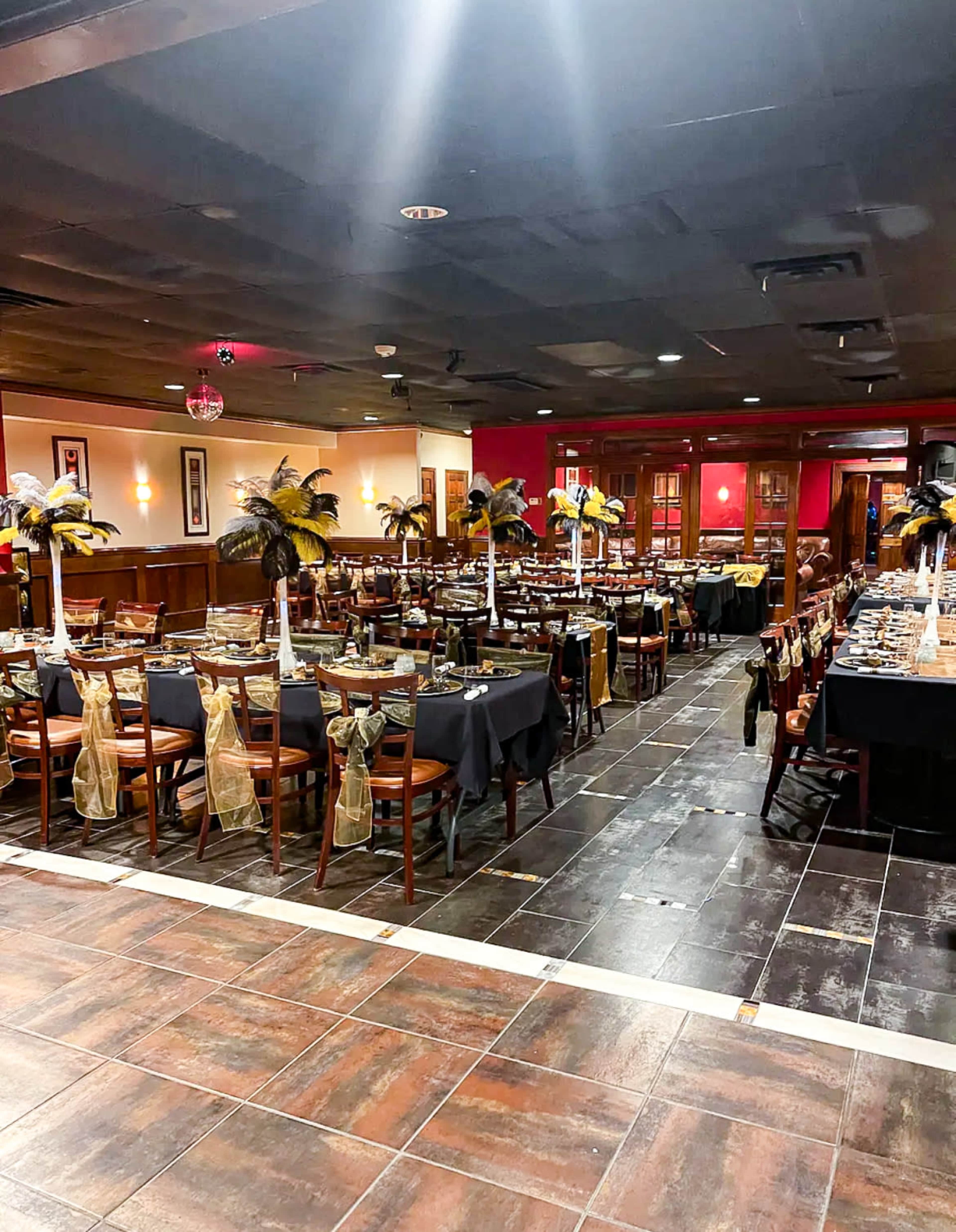 A restaurant dining area arranged with tables set for a formal event, featuring black and gold decorations and centerpieces with feathers.