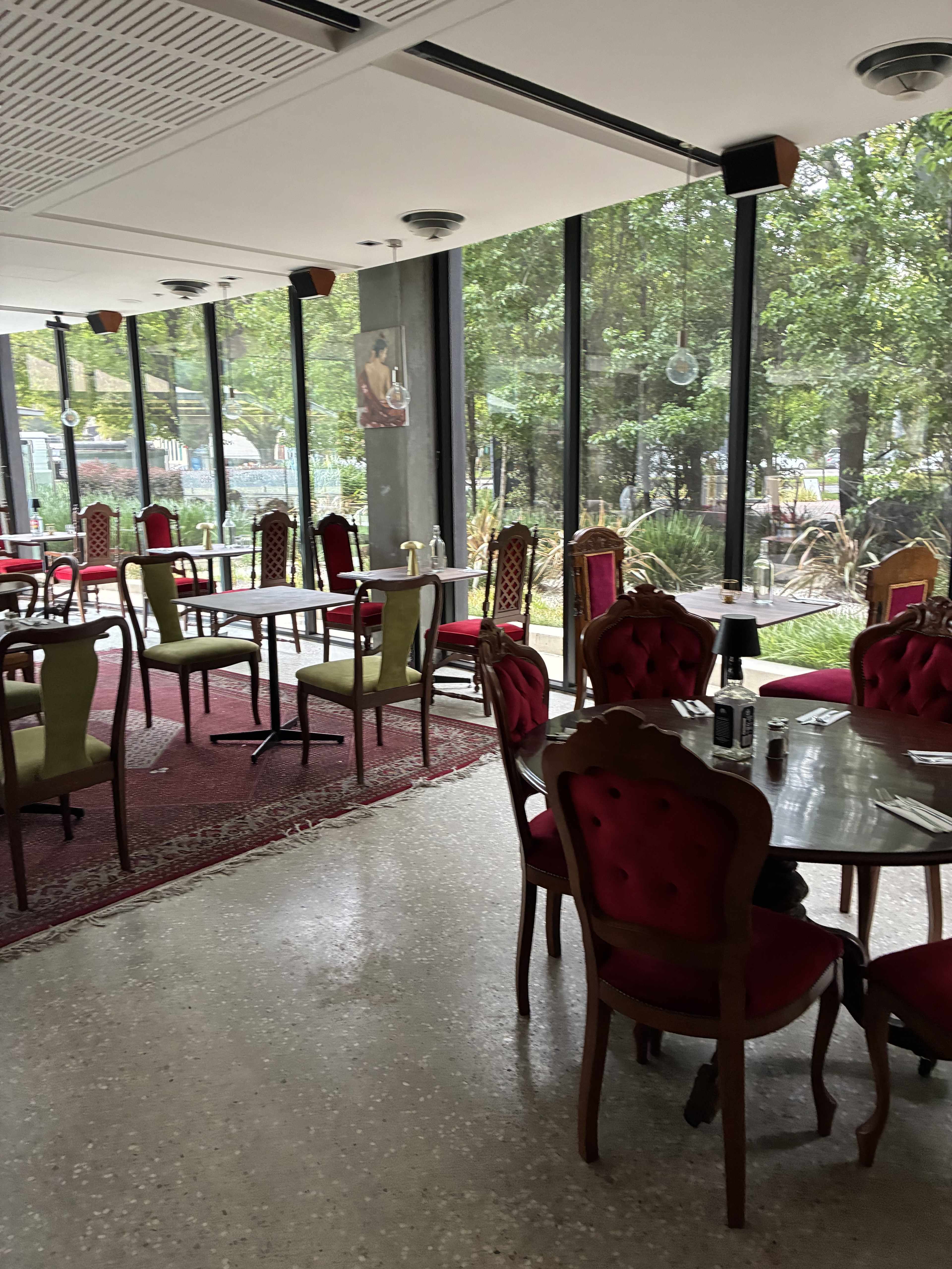 The image shows a bright room with tables and chairs arranged for dining, featuring large windows that overlook greenery outside.