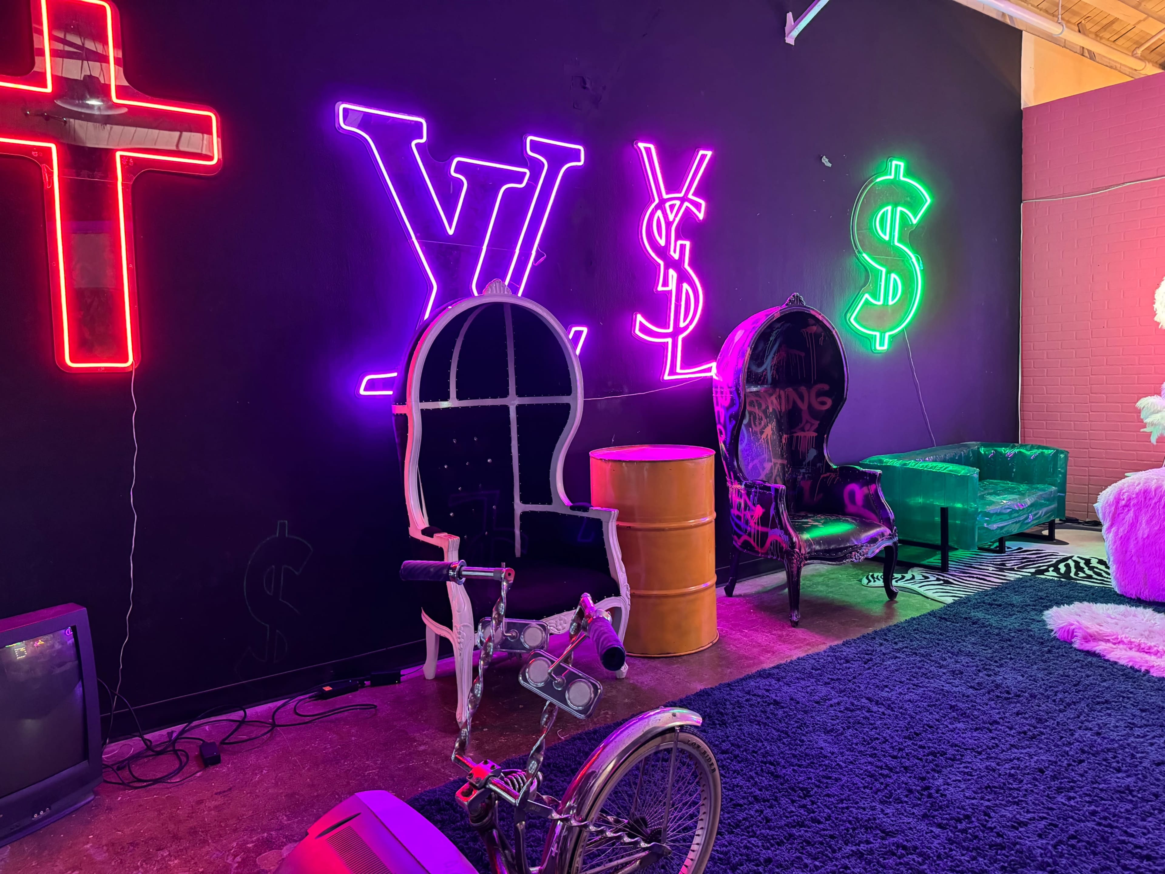 The scene features a room with neon signs of a cross, the letters "V" and "S," and dollar signs on a black wall, accompanied by decorative chairs and a vintage television.