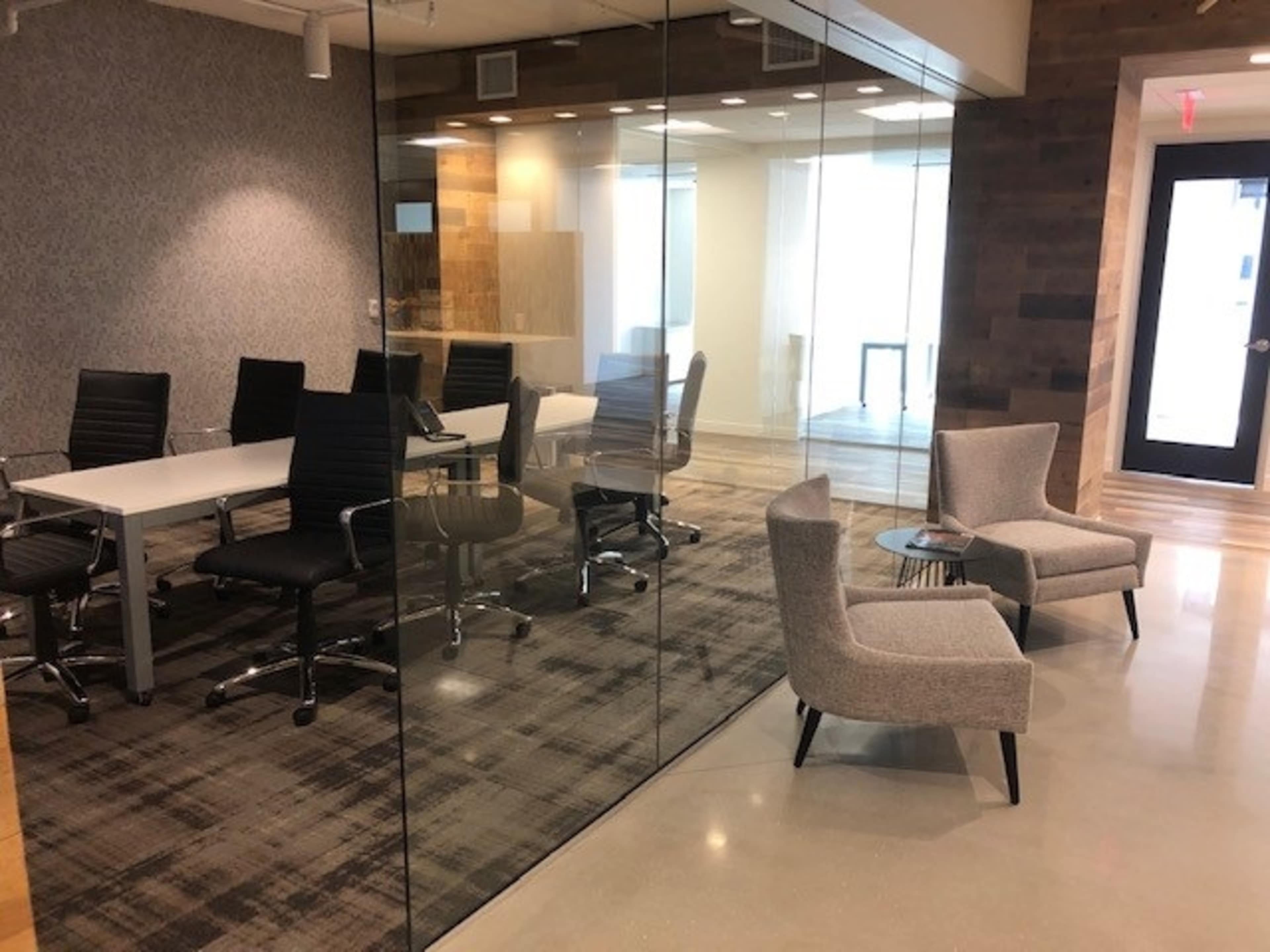 A modern office space with a glass-walled conference room featuring a table and several black chairs, alongside a seating area with two gray armchairs and a small table.
