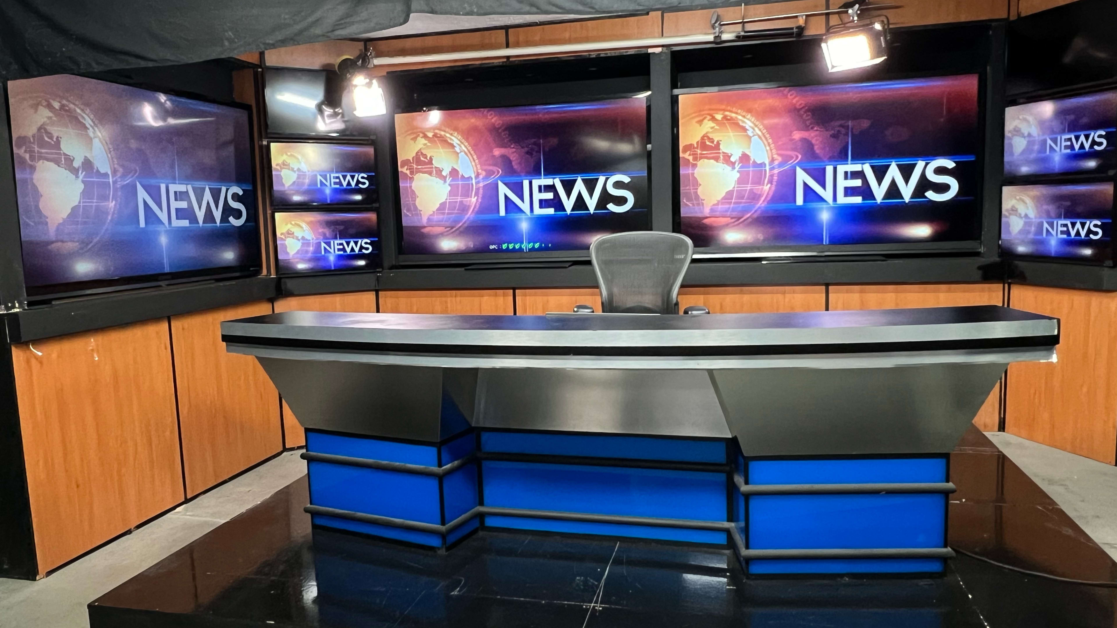 The image shows a modern television news studio with a central news desk and large screens displaying the word "NEWS."
