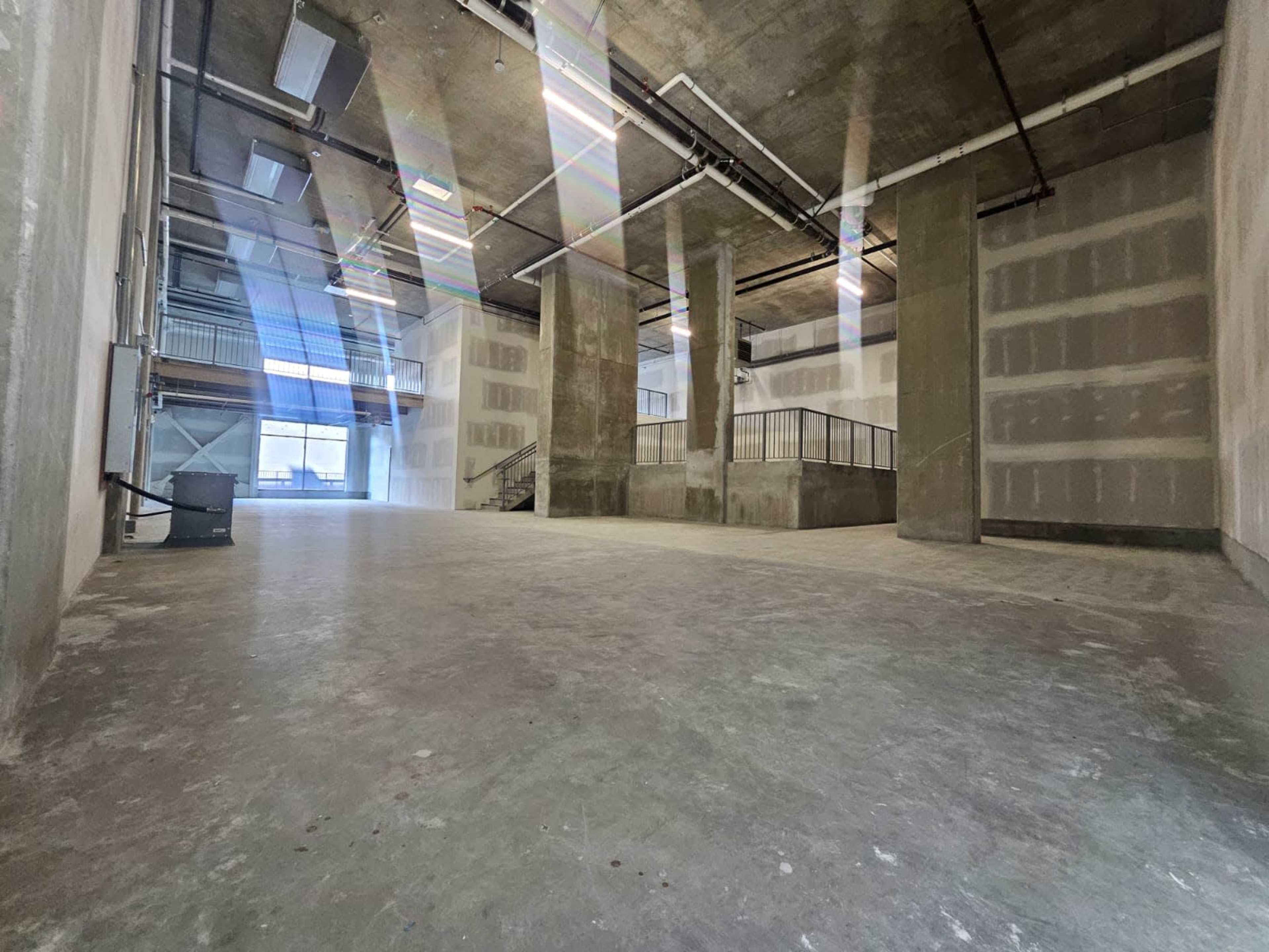 The image shows an empty industrial space with bare concrete floors and unfinished walls, featuring large windows that allow natural light to enter.