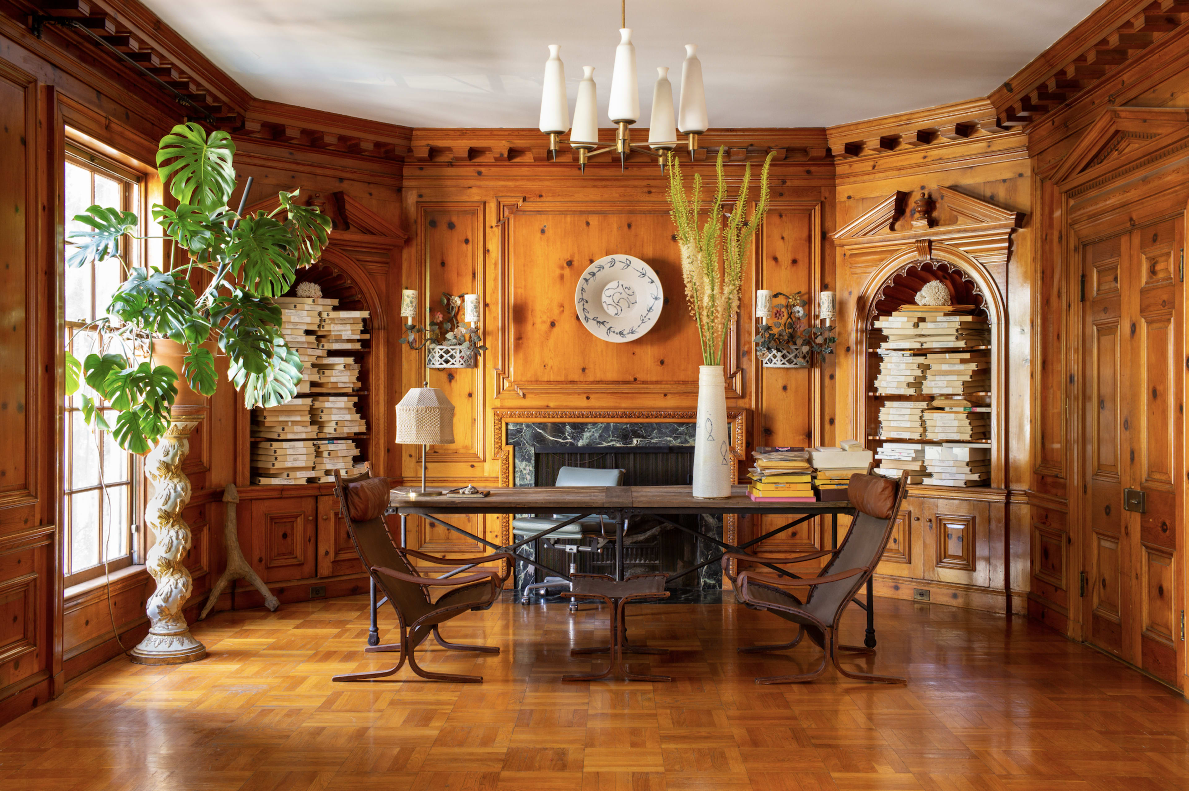 The image shows a wooden-paneled study featuring a central desk, two chairs, built-in bookshelves, and a fireplace, accompanied by decorative plants and light fixtures.
