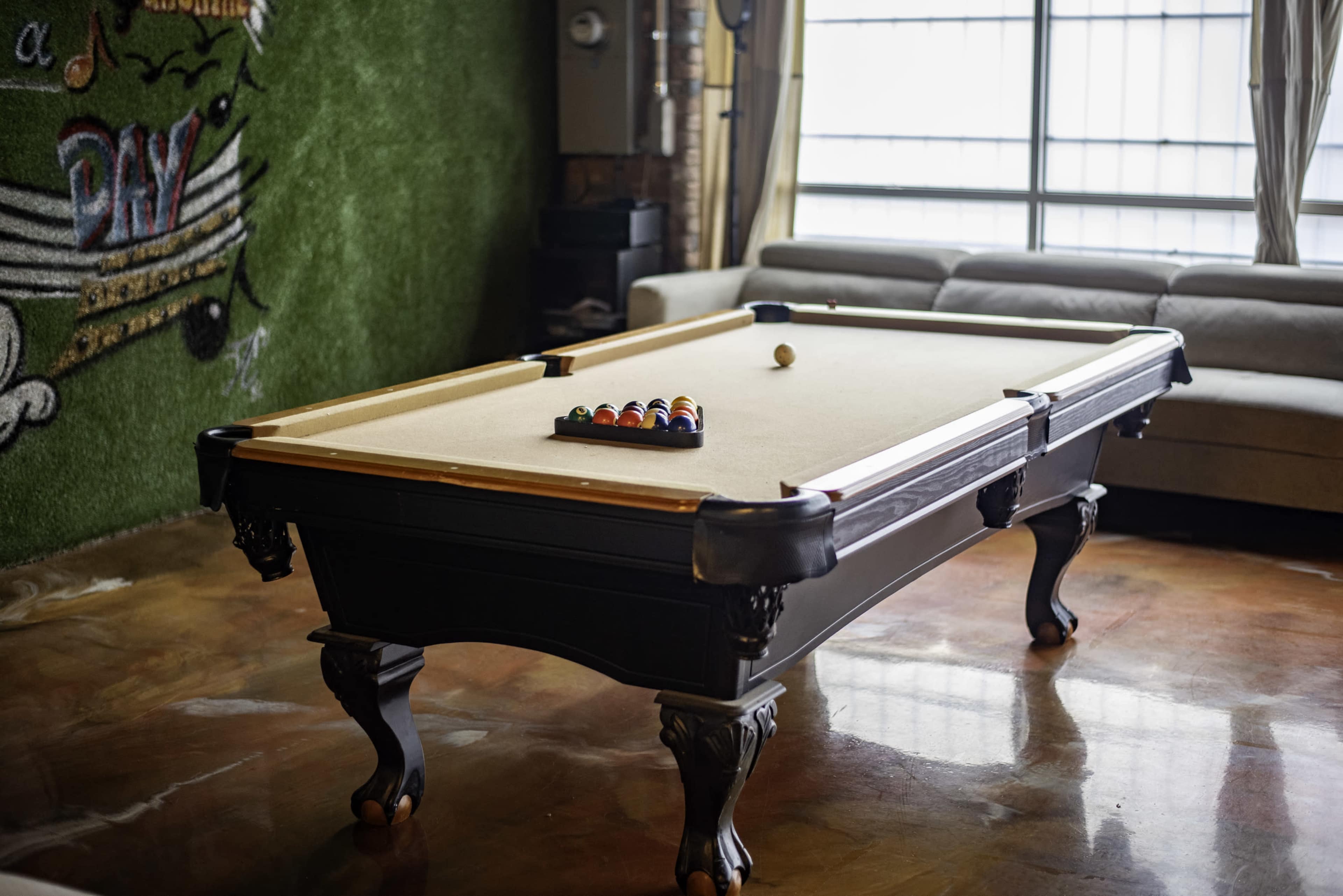 The image shows a billiards table with a set of colored balls arranged in a triangle, placed in a well-lit room with a green wall and a couch in the background.