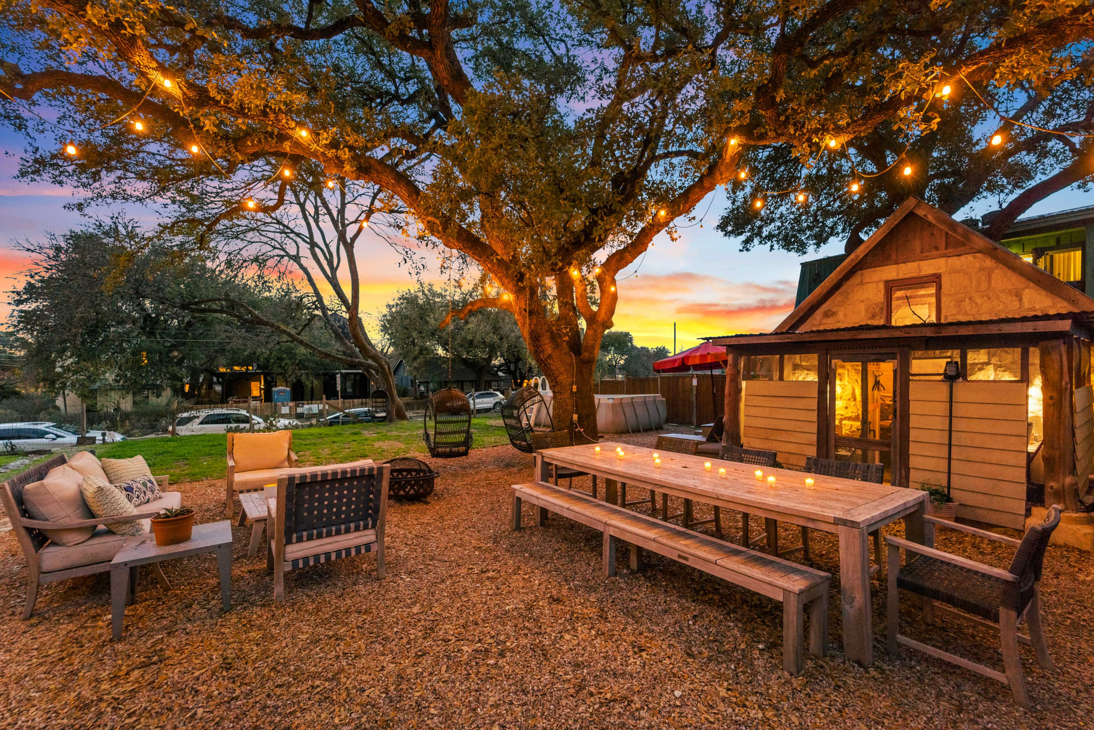 A rustic outdoor space features a long wooden dining table surrounded by benches, string lights illuminating the area, and a small shed in the background.