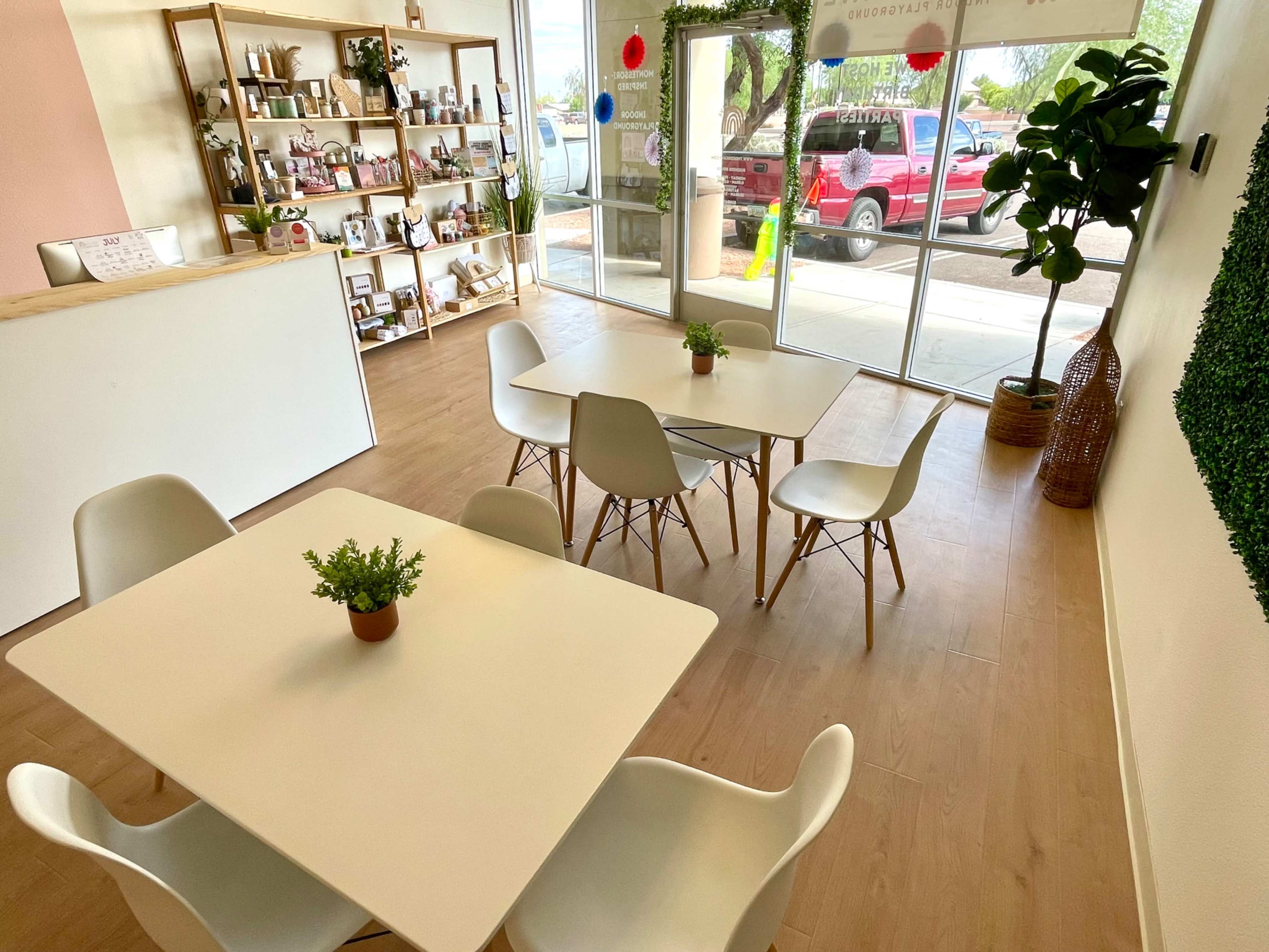 The image shows a bright interior of a café featuring wooden flooring, white tables surrounded by chairs, and decorative shelves with various items in the background.