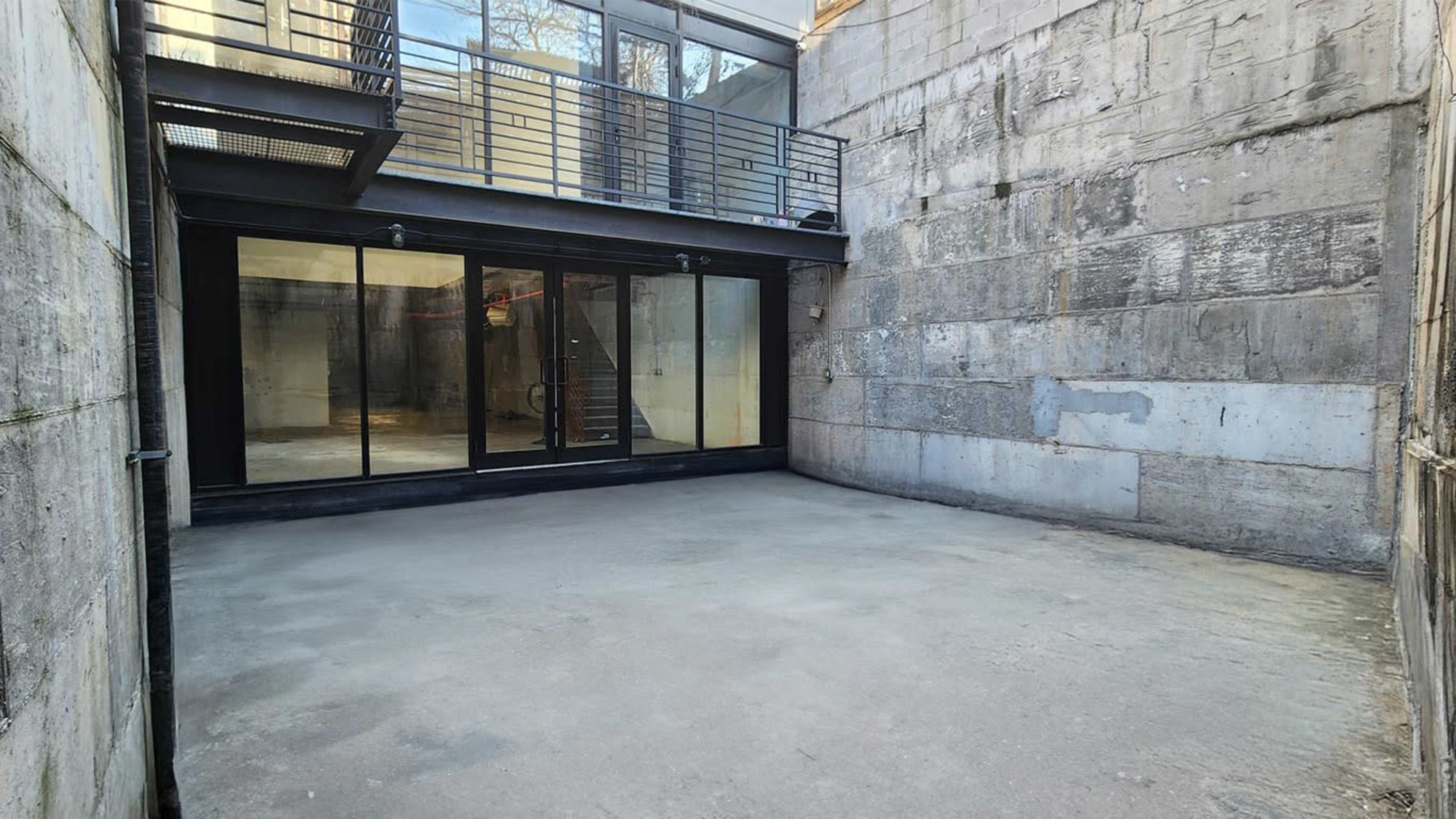 The image shows a concrete-walled courtyard with glass doors leading into an interior space, surrounded by raw, unfinished walls.