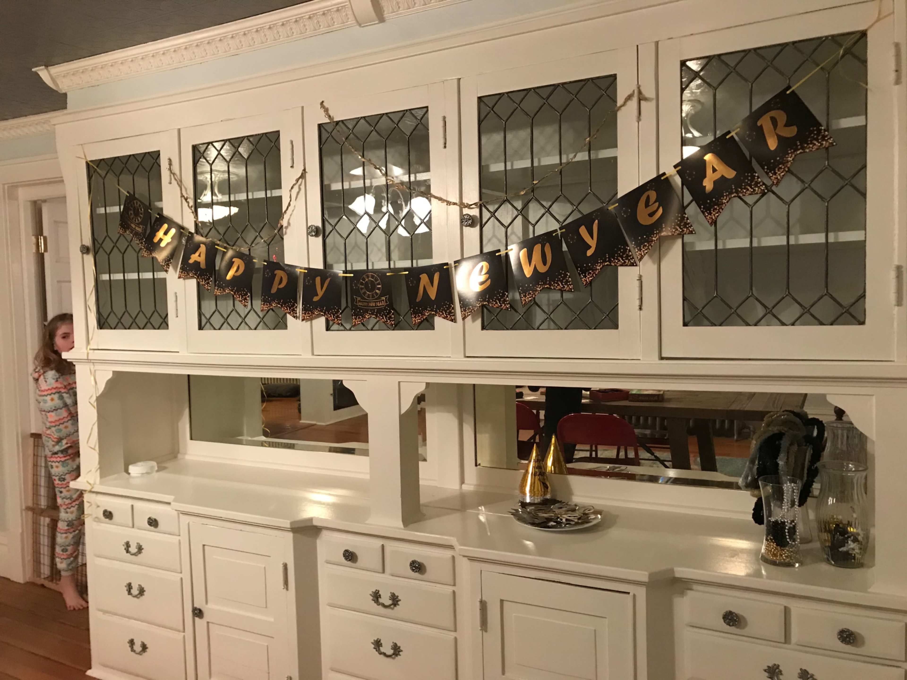 A "Happy New Year" banner hangs on a white cabinet in a room decorated for the celebration, while a child stands in colorful pajamas nearby.