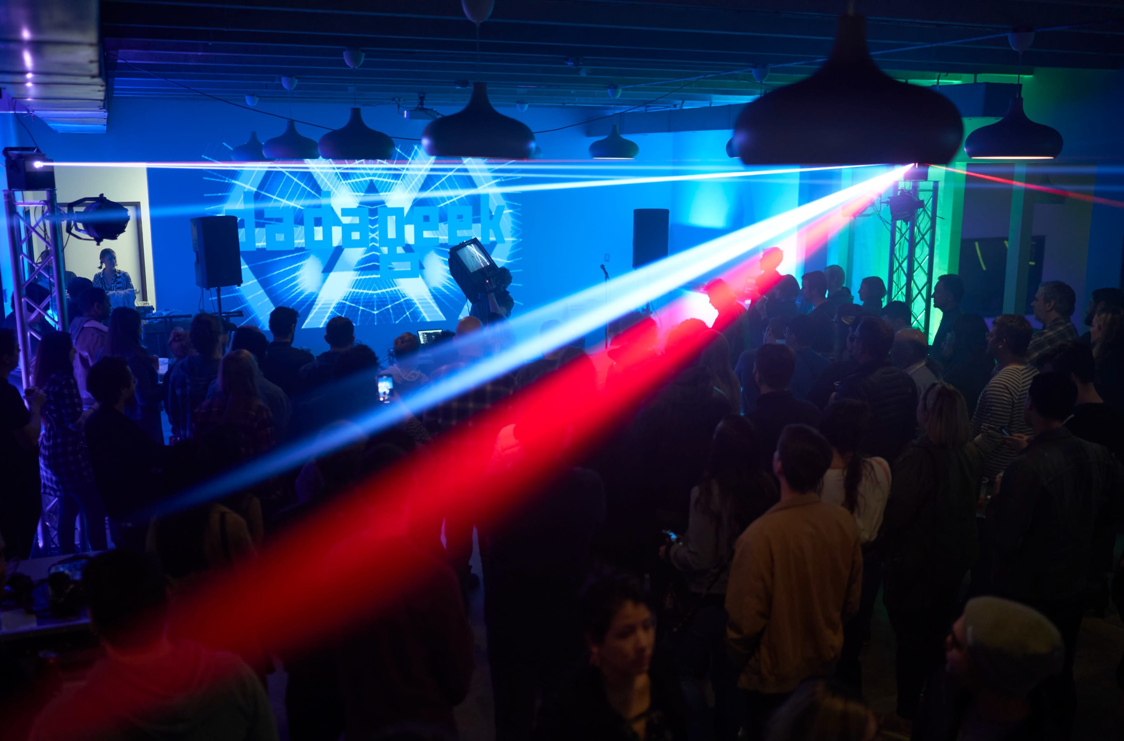 A crowd of people stands in a dimly lit venue with colorful laser lights projecting onto the wall during a live performance.
