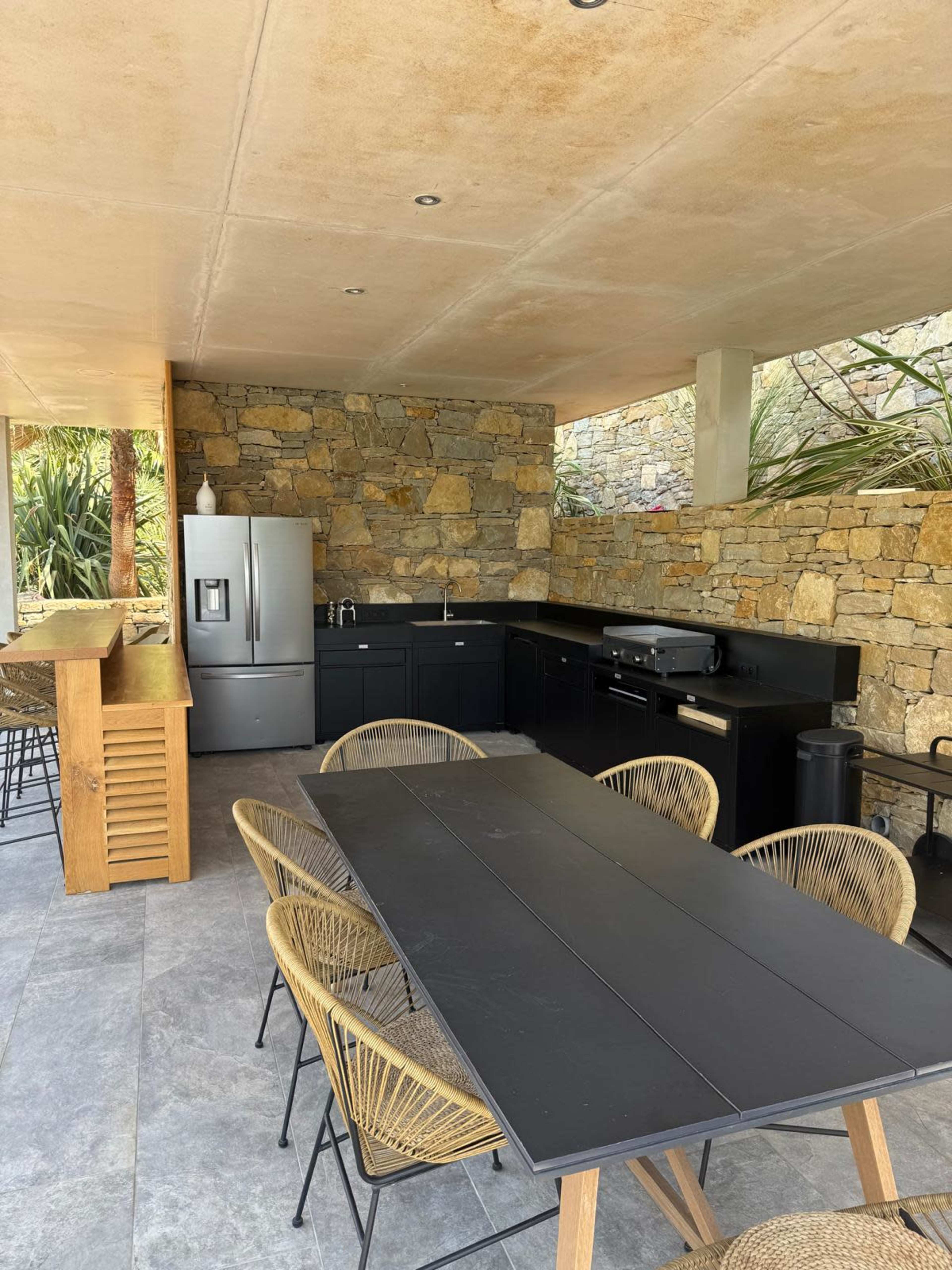 The image shows an outdoor kitchen area featuring a stone wall, black cabinetry, a silver refrigerator, and a dining table with several woven chairs.