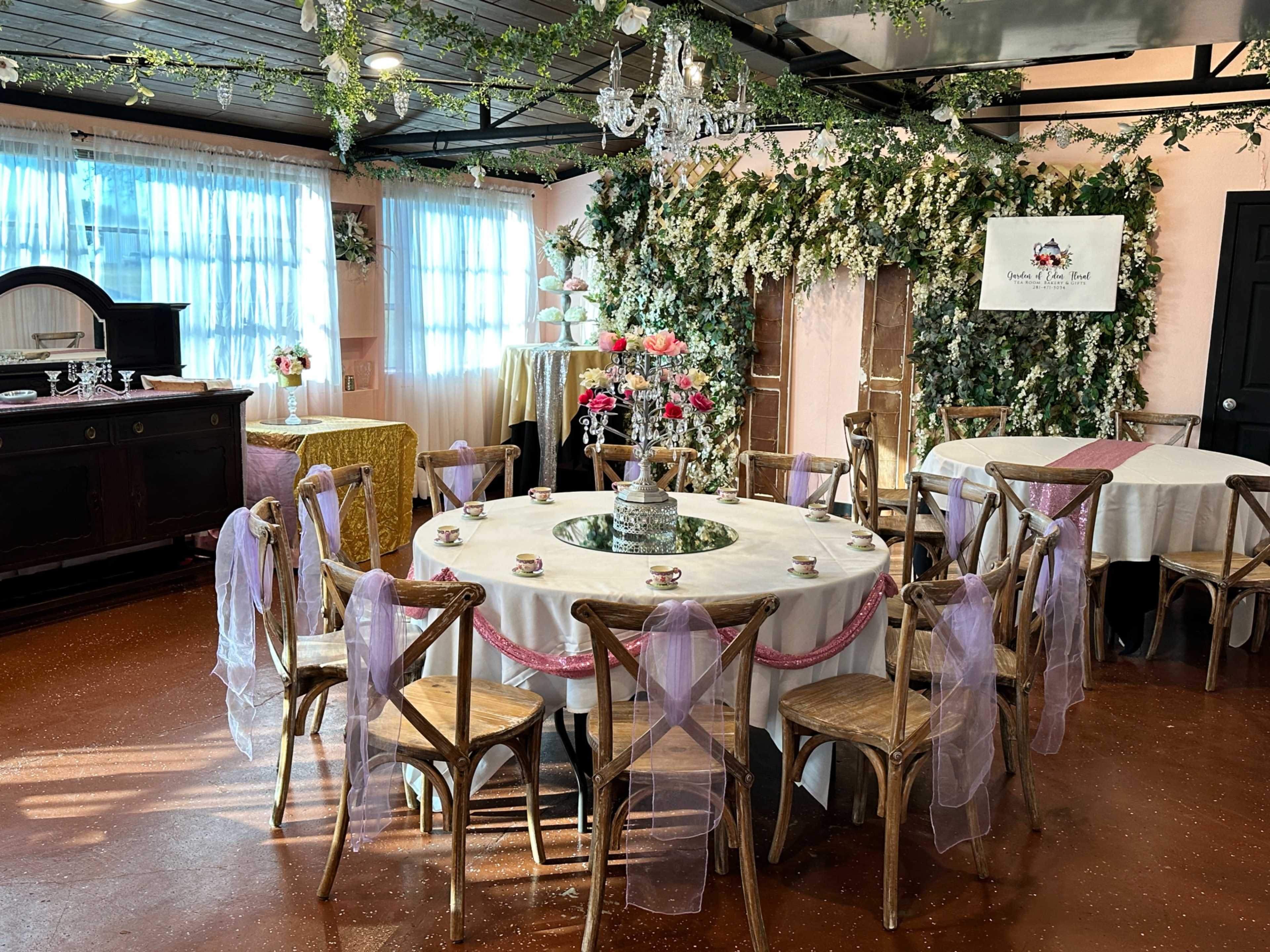 A elegantly decorated event space features a round table set with floral arrangements and surrounded by chairs with pastel ribbons, against a backdrop of greenery and soft drapery.