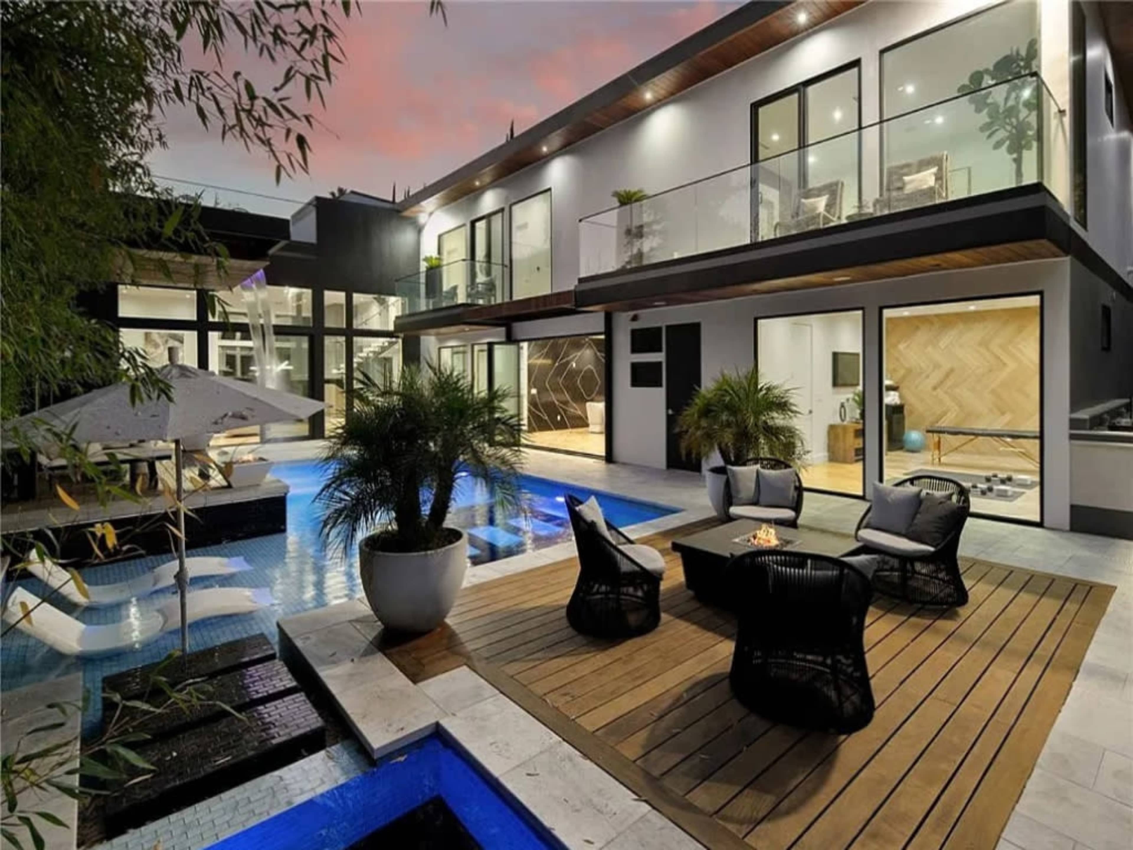 The image shows a modern home with large glass windows surrounding a swimming pool and a wooden deck featuring seating areas and a fire pit.