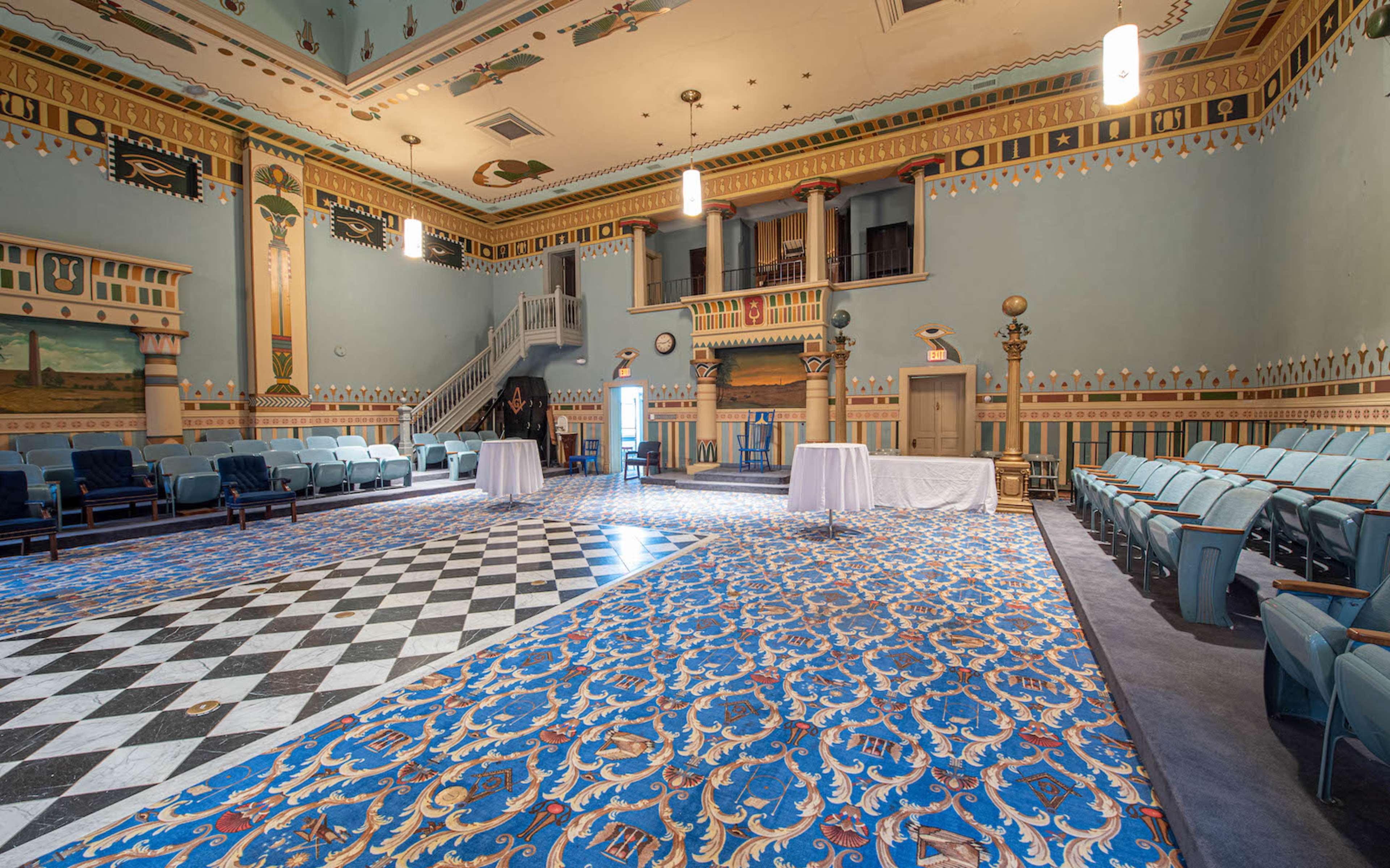 A spacious, ornate hall with a patterned blue carpet, checkerboard flooring, and seating arranged in rows facing a stage.