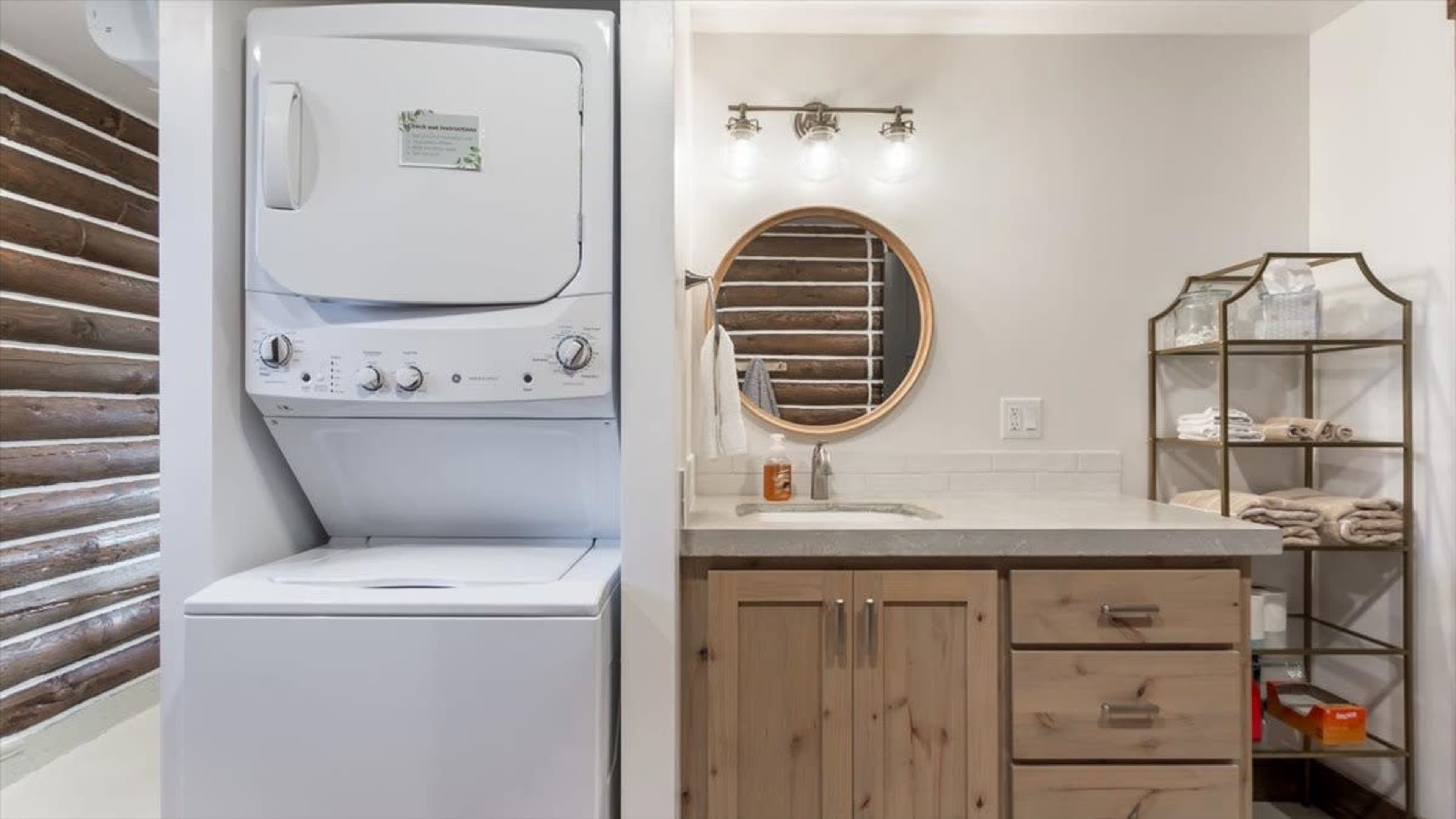 The image shows a laundry area with a stacked washer and dryer on the left and a bathroom vanity with a round mirror and shelving on the right.