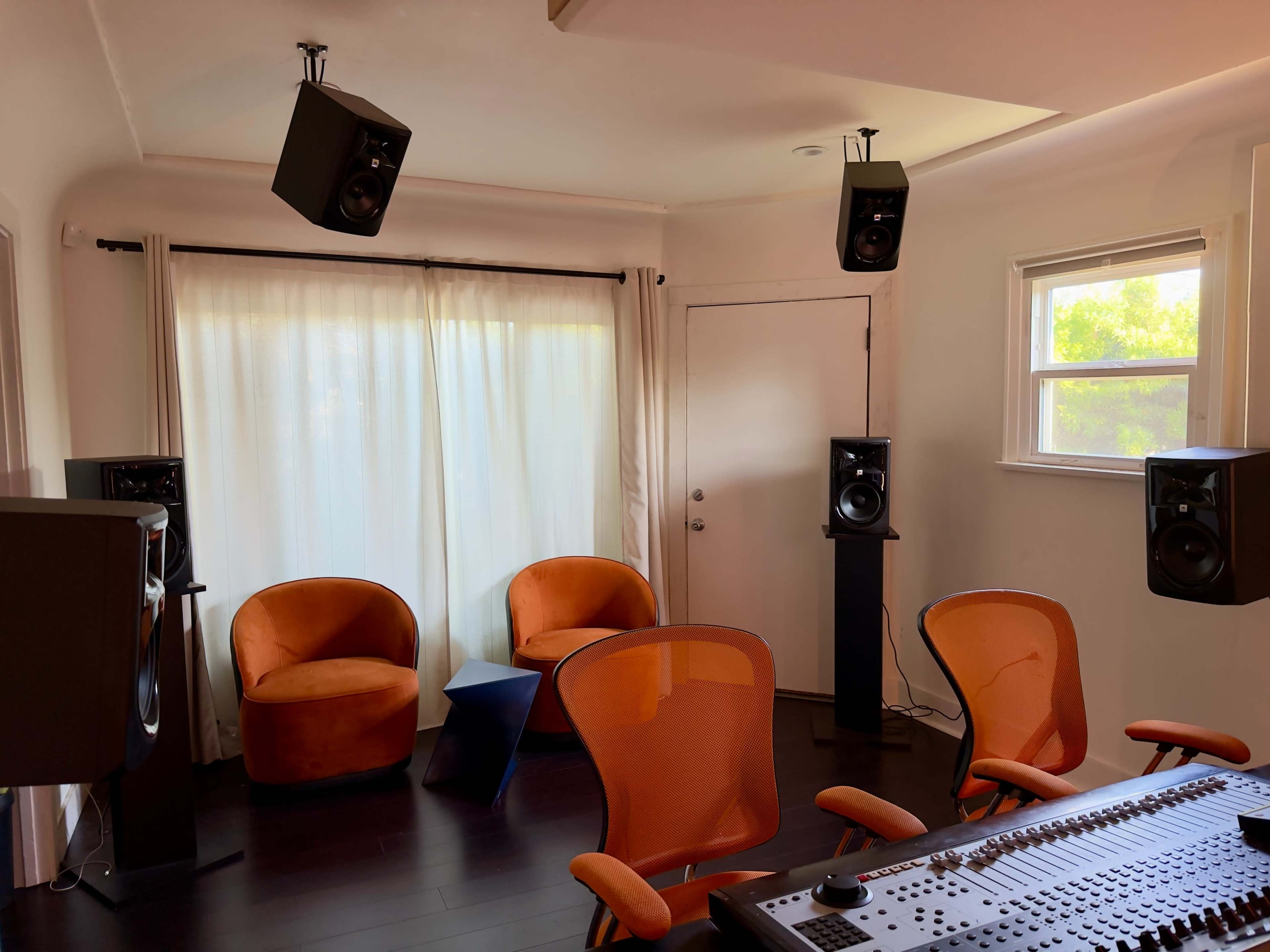 The image shows a music studio with two orange chairs, a blue table, and several speakers positioned around the room.