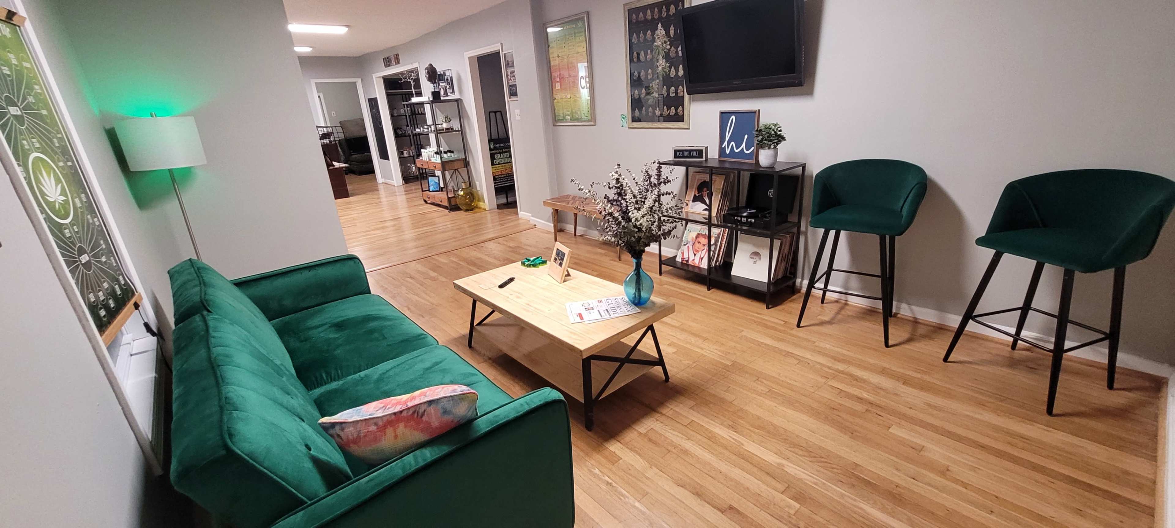 A green couch and a wooden coffee table are positioned in a living area with light wooden floors, flanked by two green bar stools and decorated with wall art and shelves.