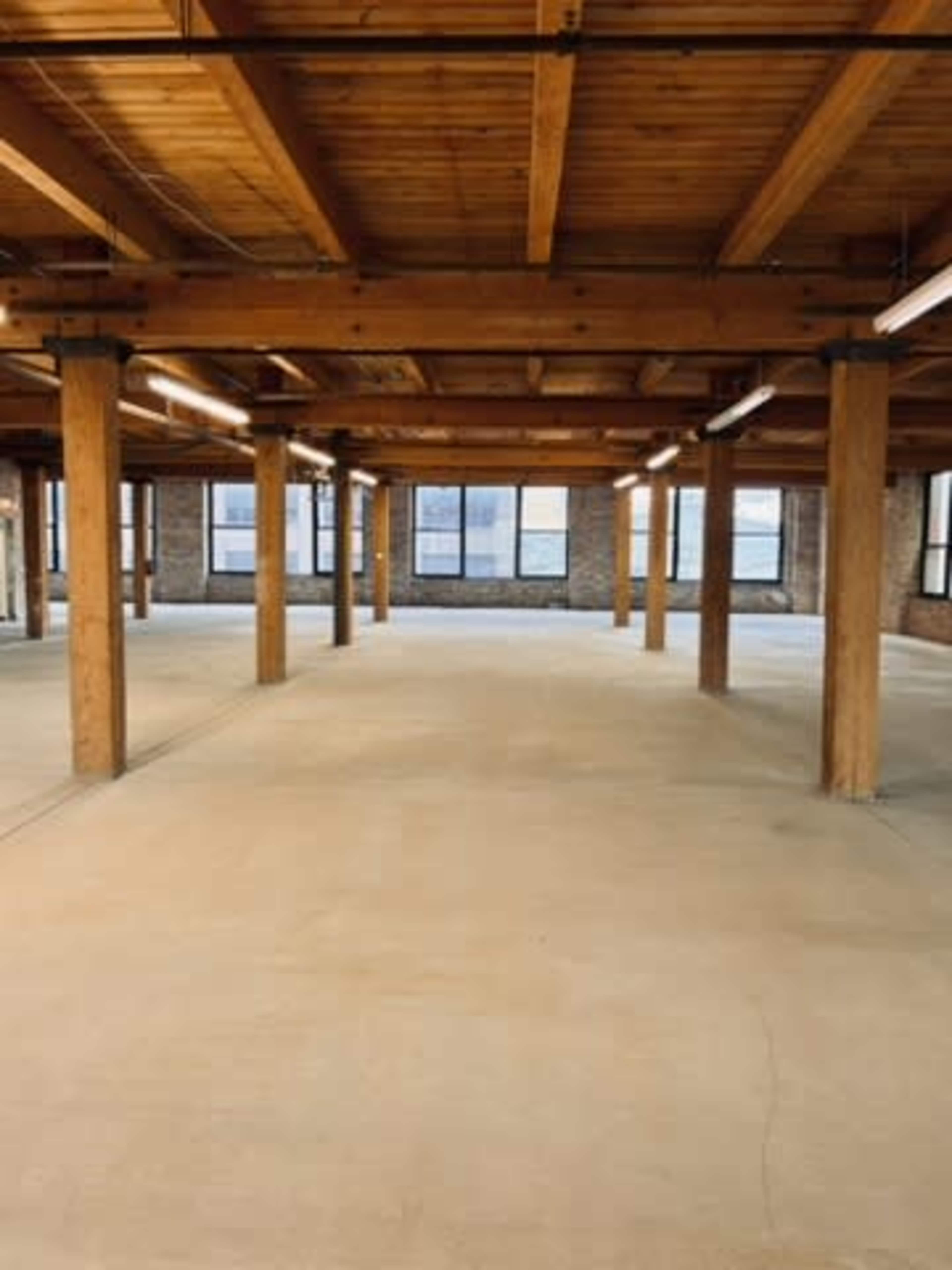 An empty industrial space features exposed wooden beams and columns, with large windows allowing natural light.